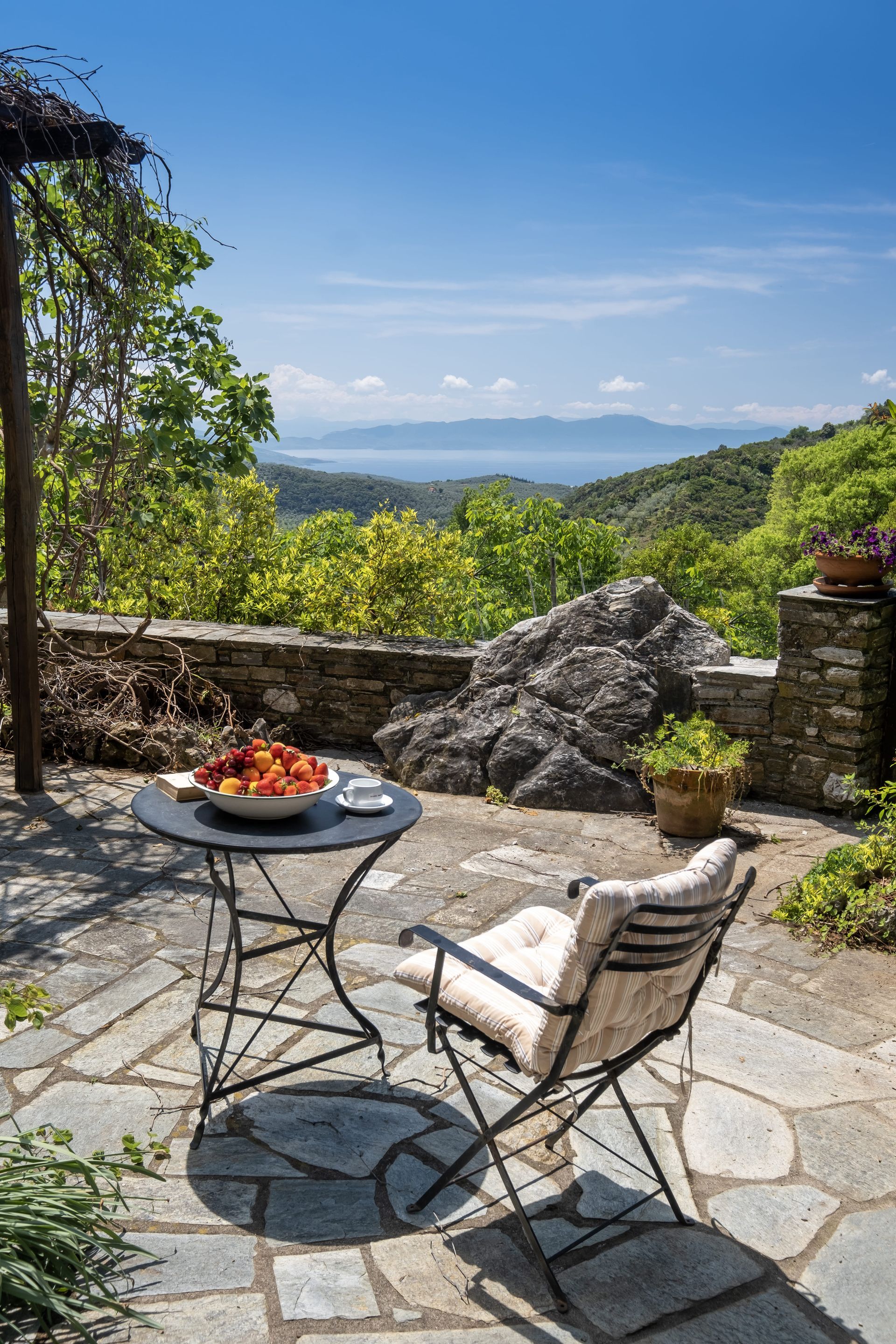 Patio with table and chair overlooking a scenic view. Fruit bowl on table, blue sky.