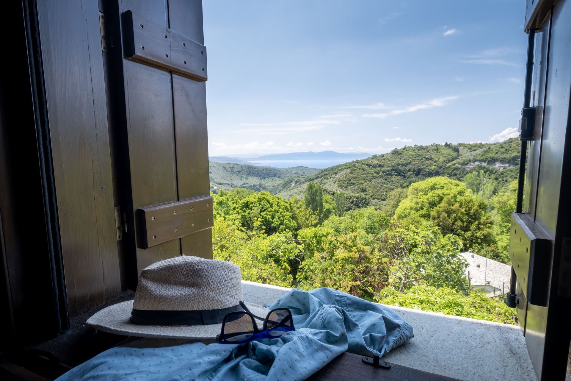 A straw hat, sunglasses, and blue cloth on a windowsill overlooking a green, hilly landscape and blue sky.