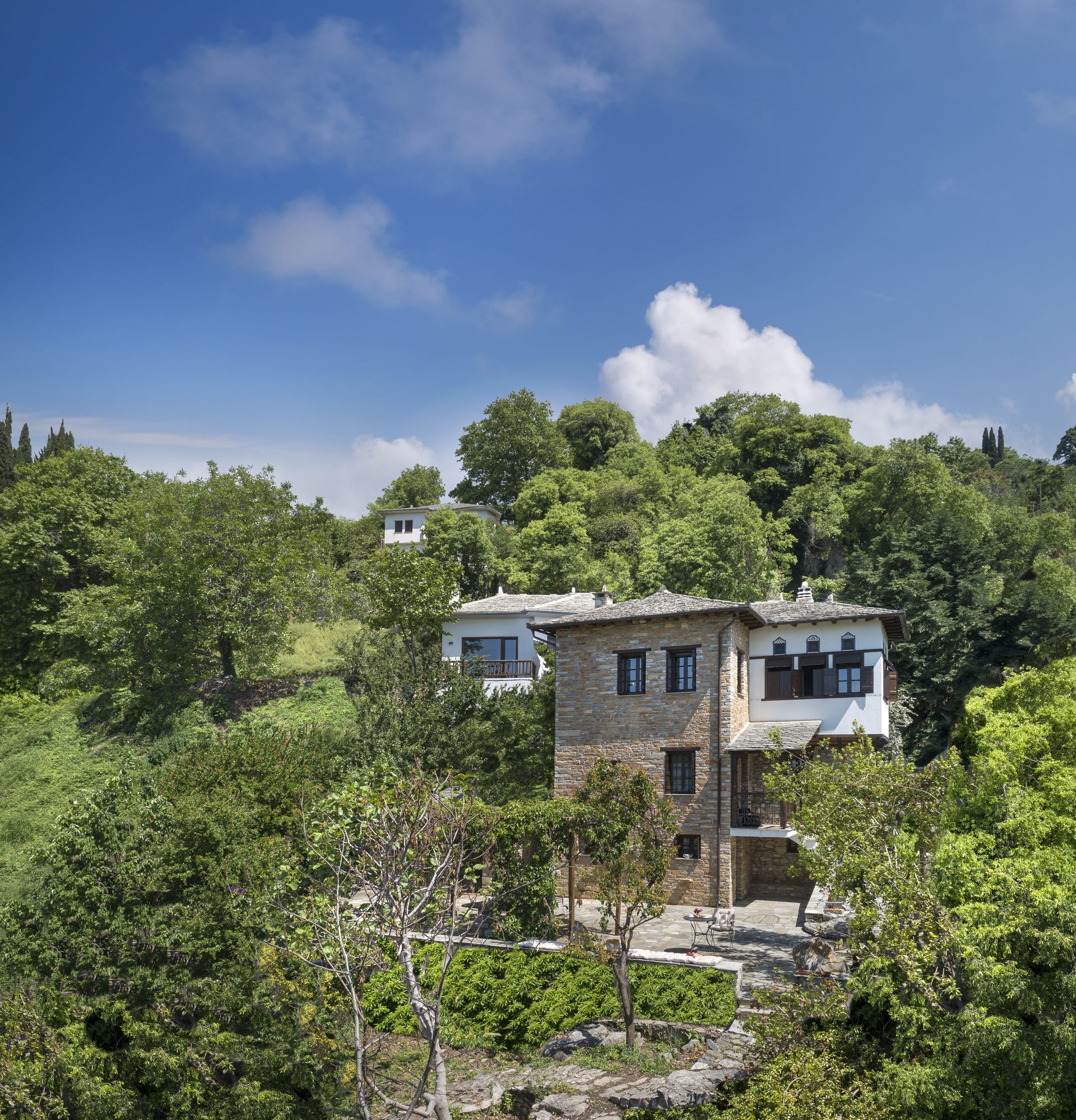 Stone buildings nestled amongst green trees under a blue sky with clouds.