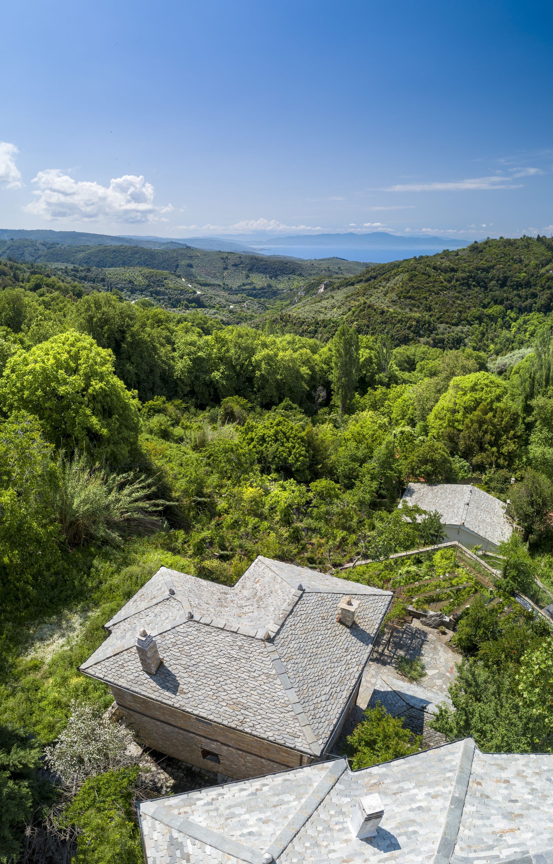 Stone-roofed buildings nestled in a lush green valley, with a glimpse of blue sea in the distance under a clear sky.