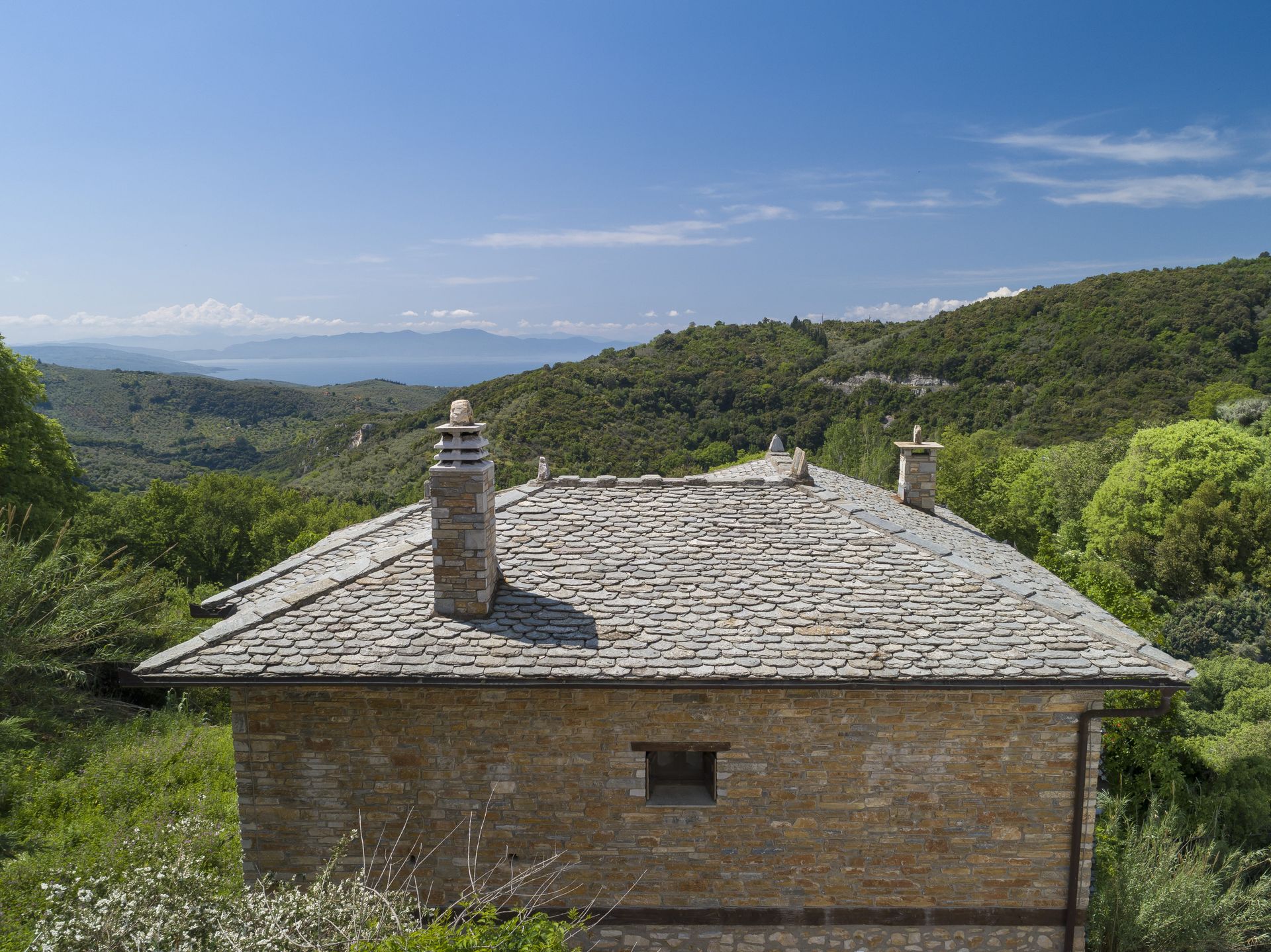 Stone house with slate roof and chimneys, nestled in a green hillside with a distant view of the sea and sky.