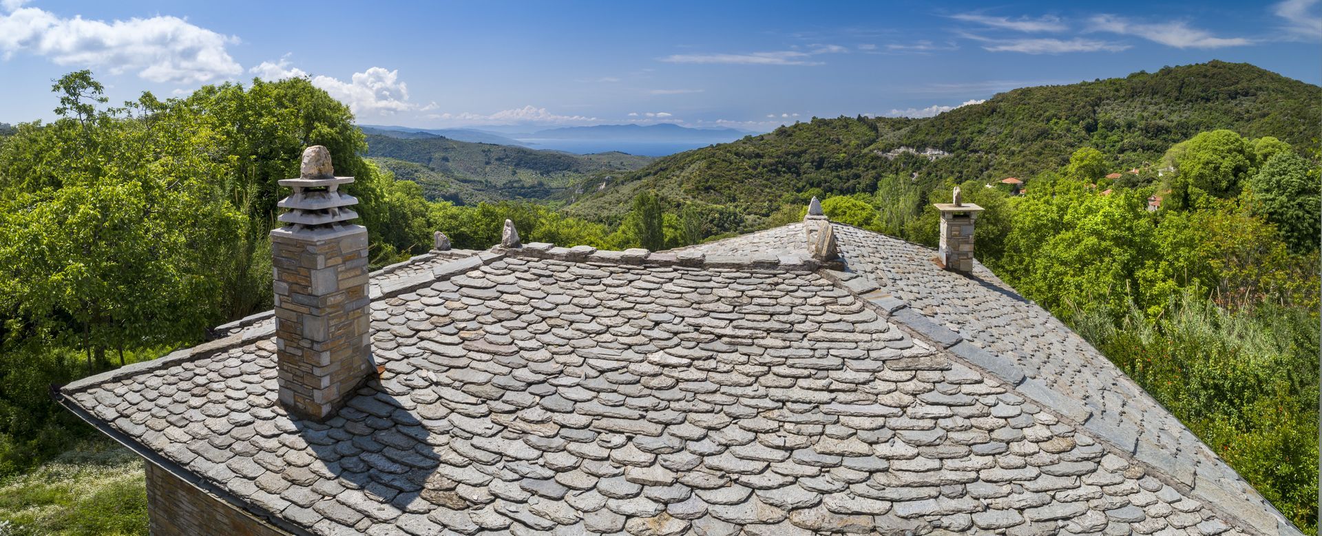 Stone roof with chimneys, overlooking a valley with trees and water under a blue sky.