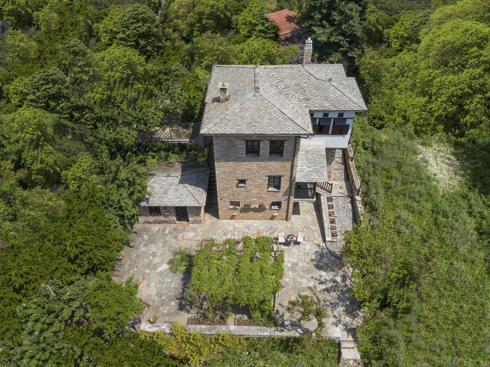 Stone house with a slate roof, surrounded by trees and a paved terrace.