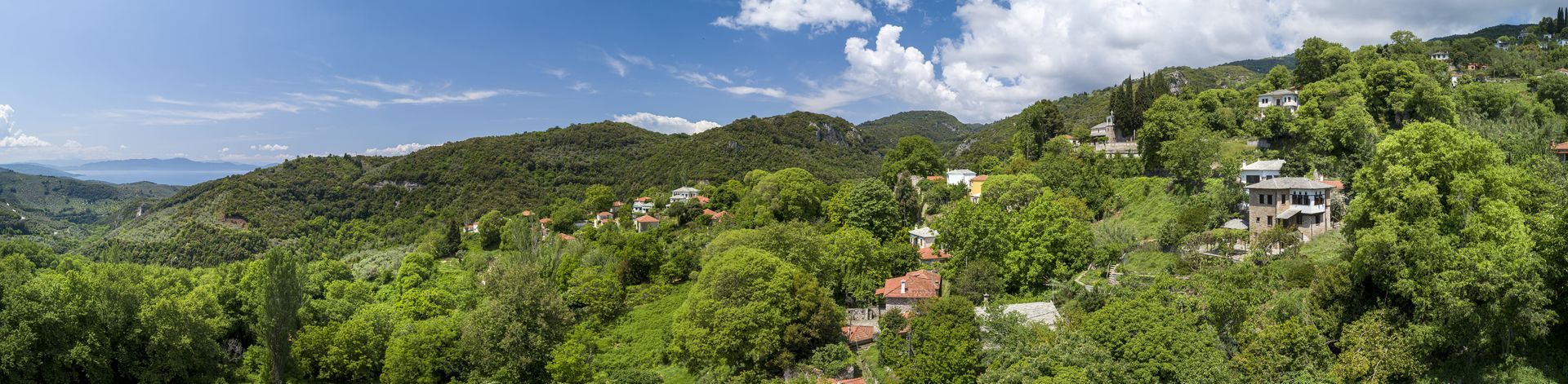 Panoramic view of a lush green hillside dotted with buildings under a blue sky with clouds.