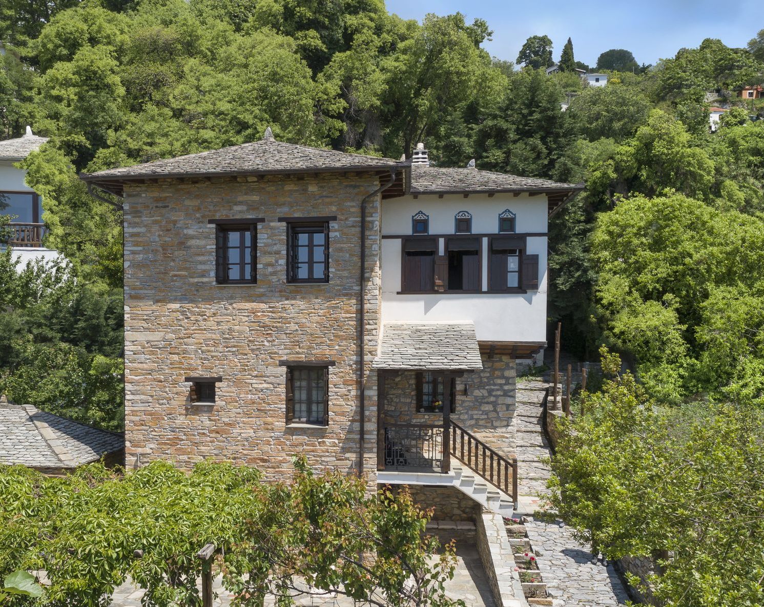 Stone house with white balcony, wooden shutters, surrounded by lush green trees.