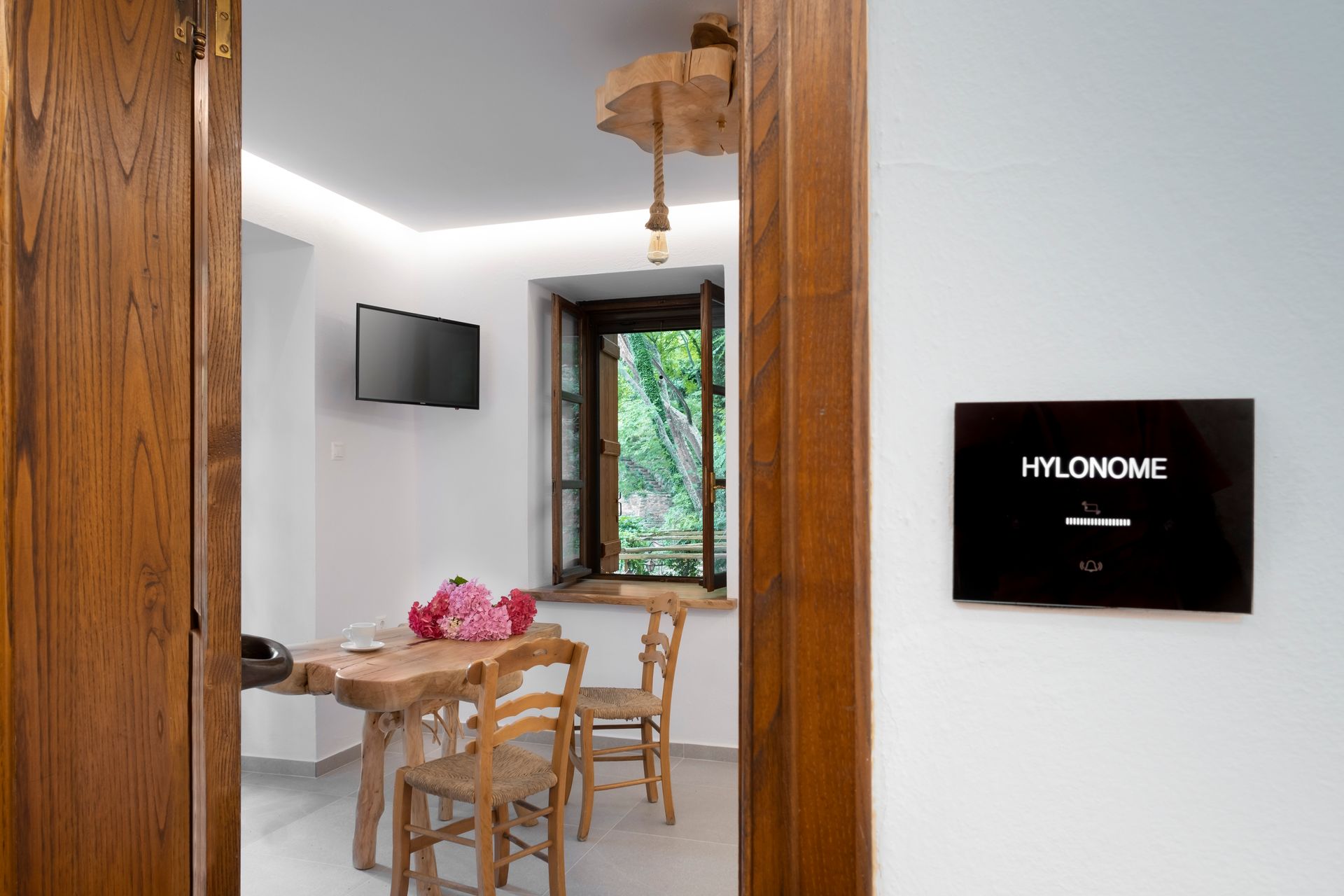 Wooden doorway into a room with a rustic table, chairs, and window overlooking trees. Sign on wall says HYLOMOME.