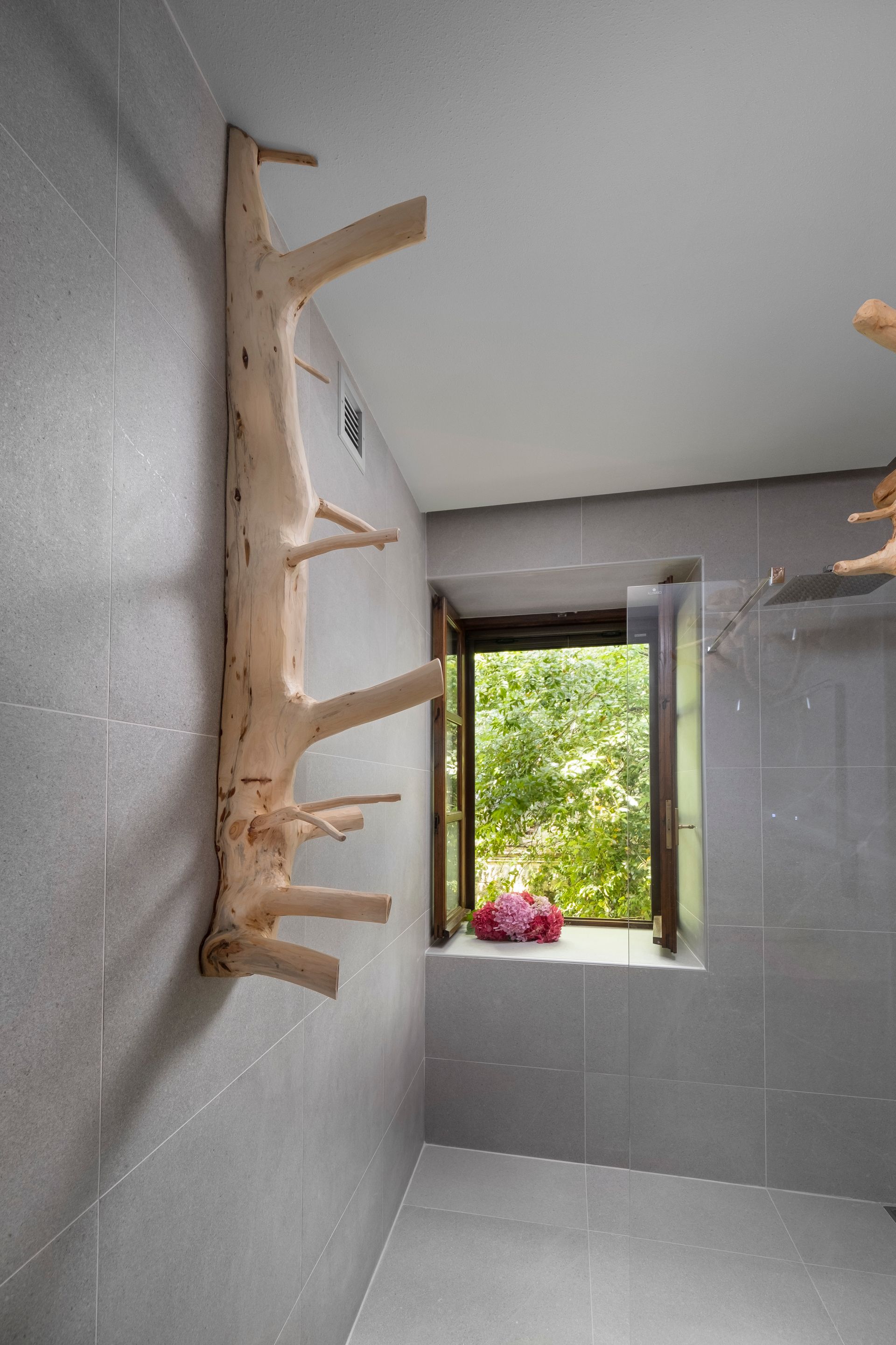 A tree branch wall rack in a gray-tiled bathroom next to a window with a view of greenery.