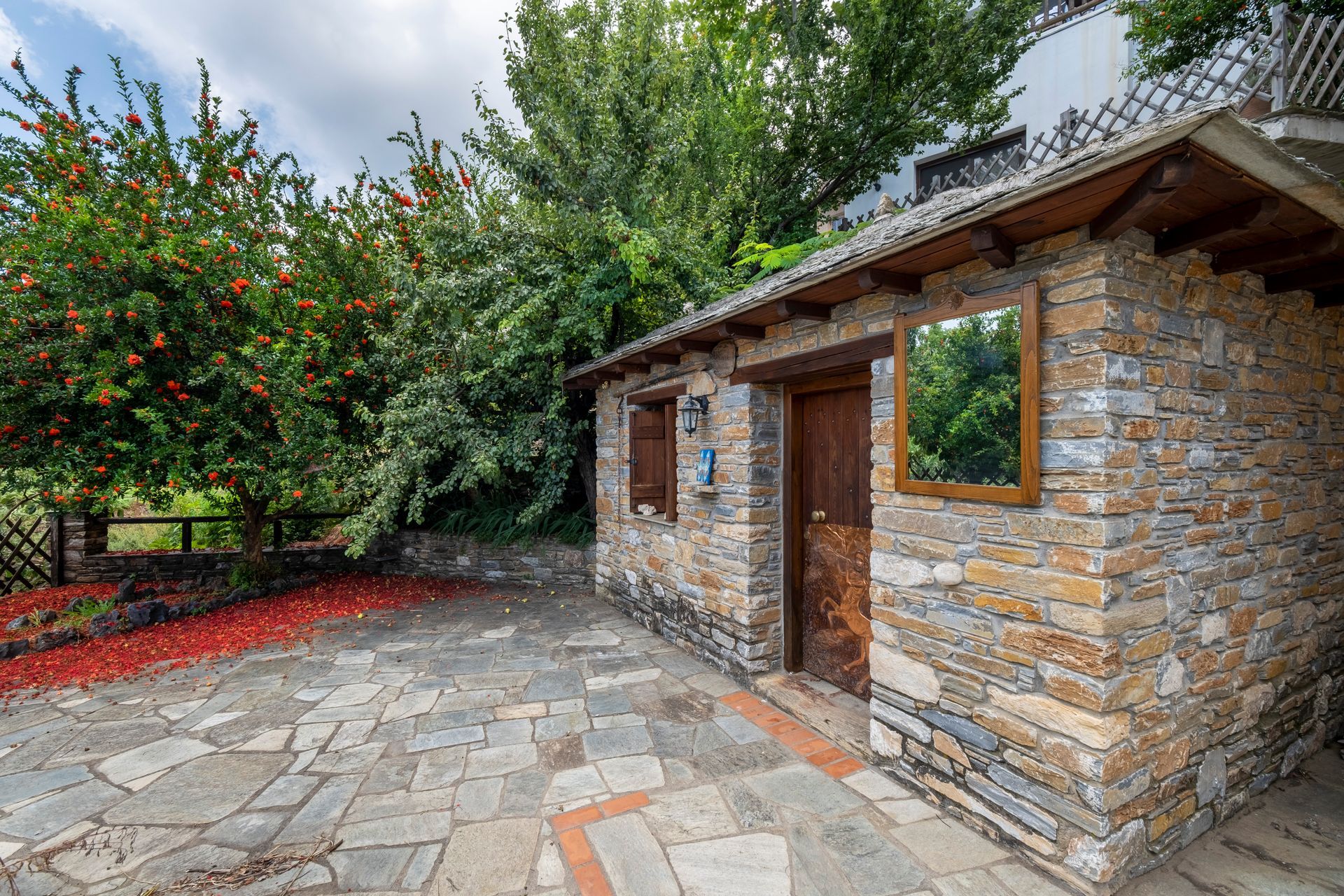 Stone building with wooden door and window, paved patio, red mulch, and leafy trees.