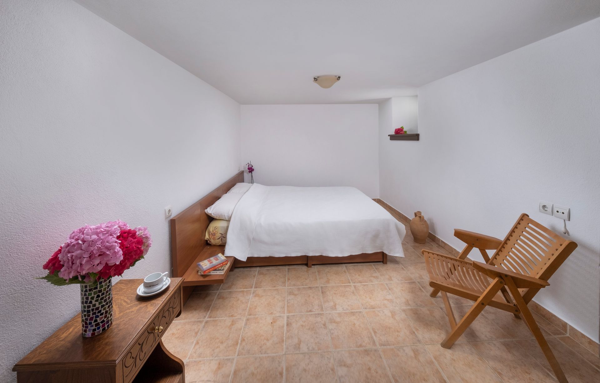 Bedroom with a bed, wooden furniture, and flowers on a table. The walls are white and the floor is tiled.
