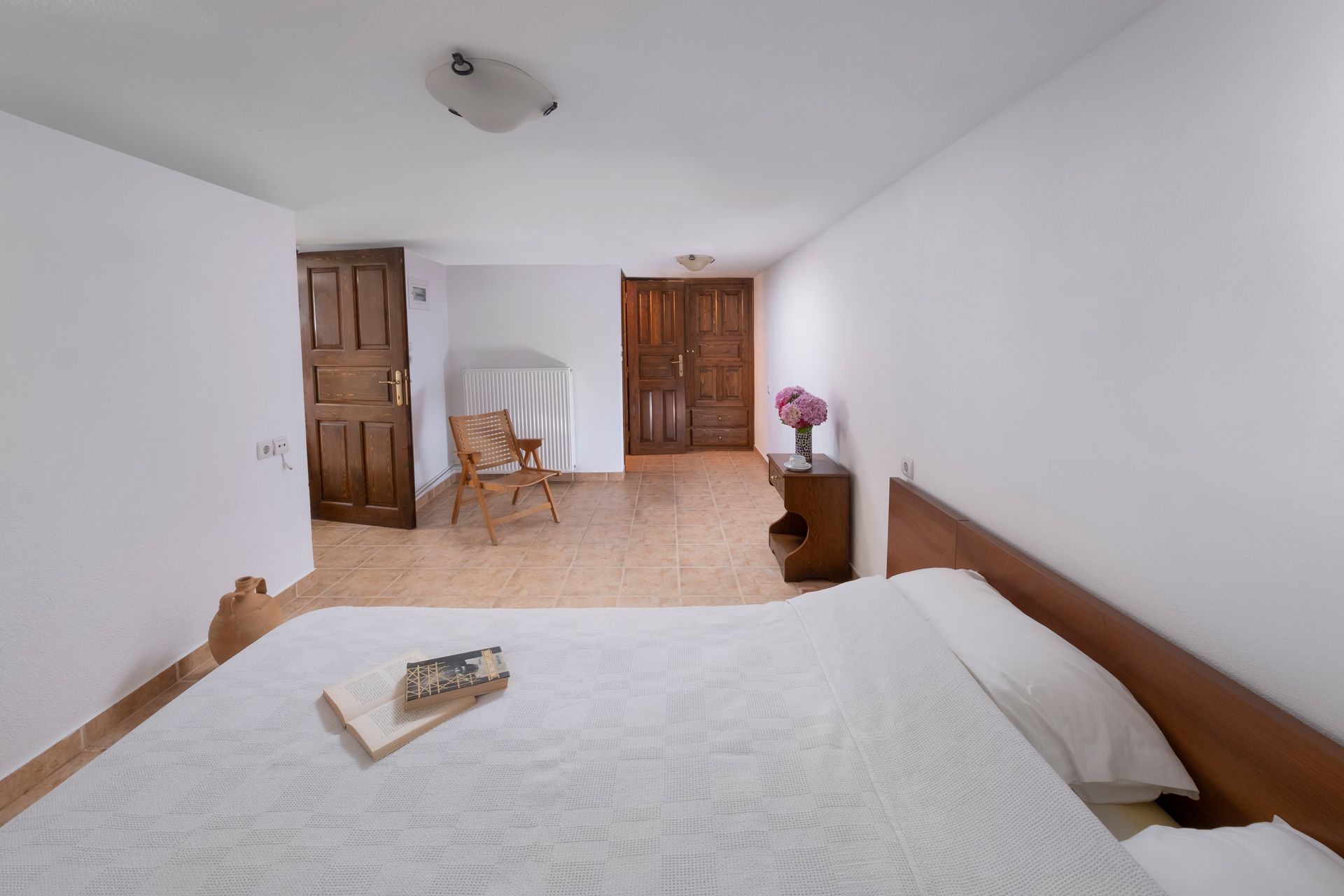 Bedroom with a bed, wooden doors, a chair, and a table with flowers. White walls and tiled floor.