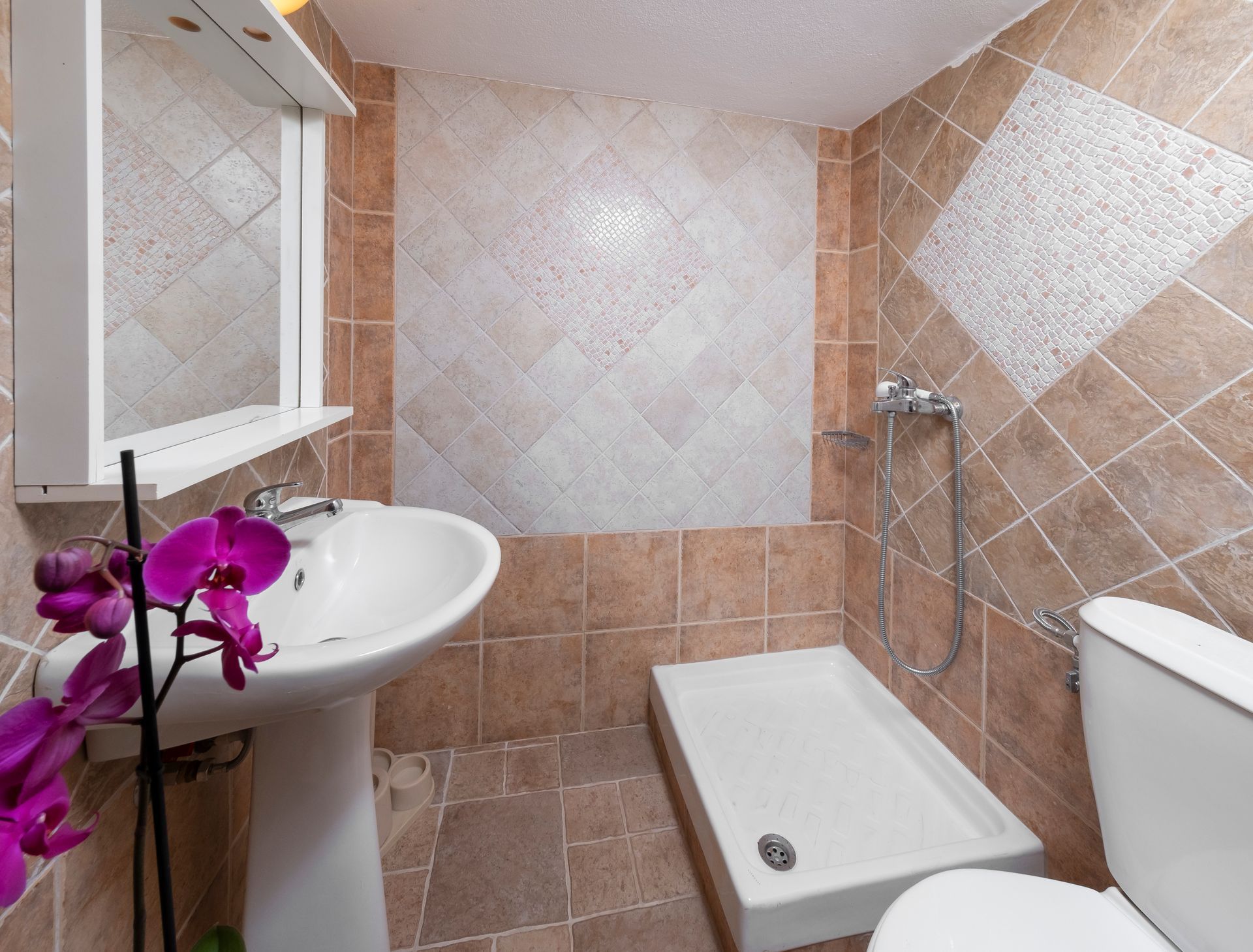 Small bathroom with pedestal sink, toilet, shower, and orchid. Brown and white tile.
