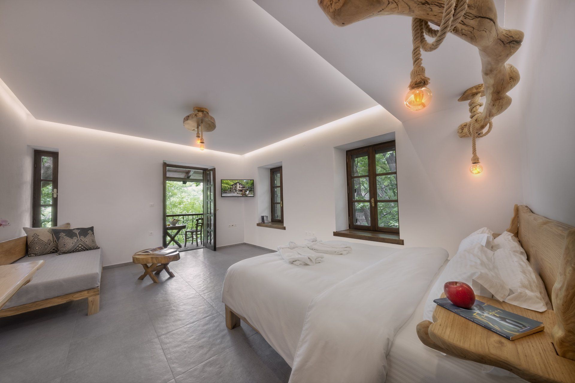 Bright hotel room with a bed, sofa, and balcony access. Wooden decor, white walls, and warm lighting.