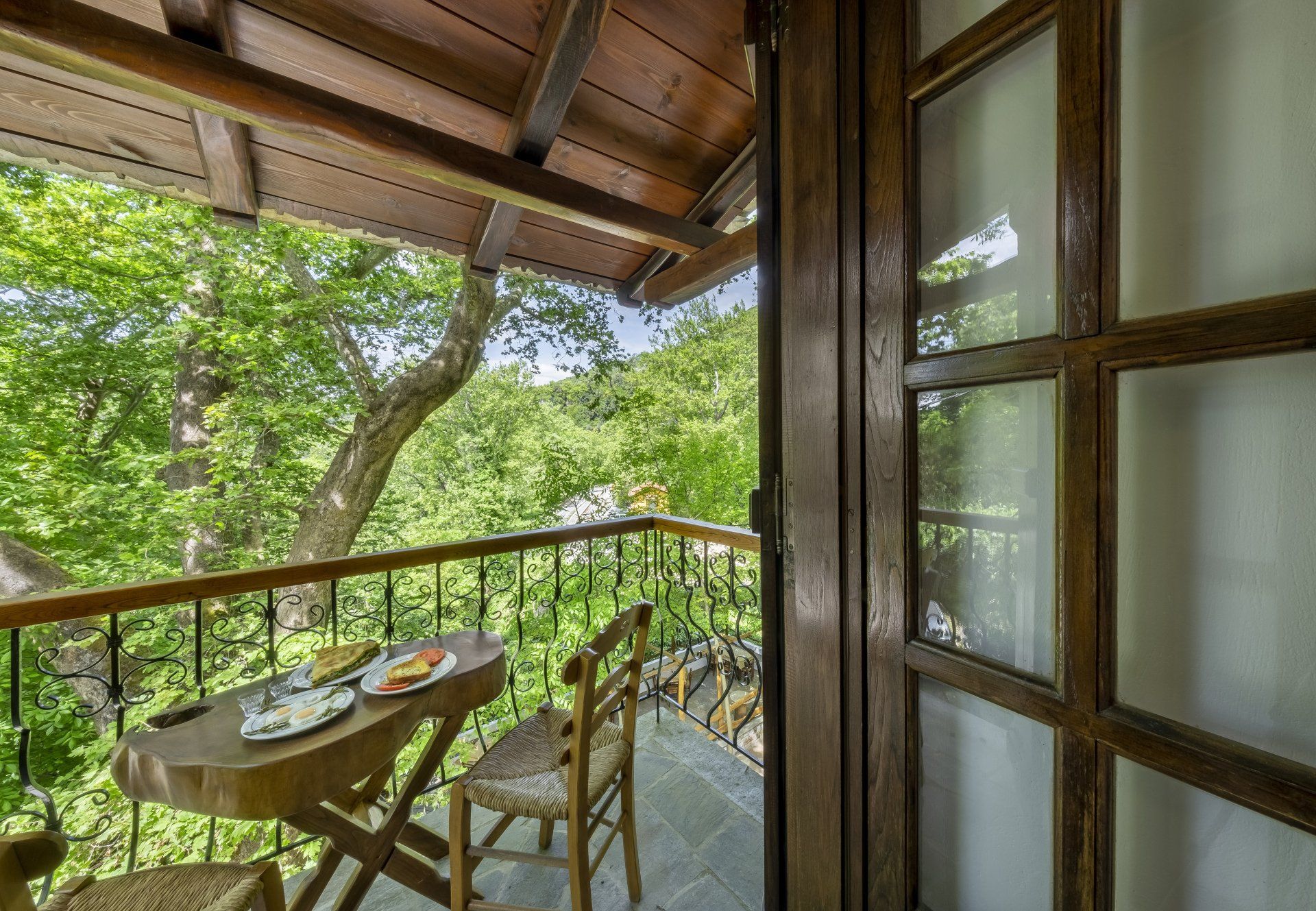 Balcony with table set for food, overlooking lush green trees. Wooden door and railing.