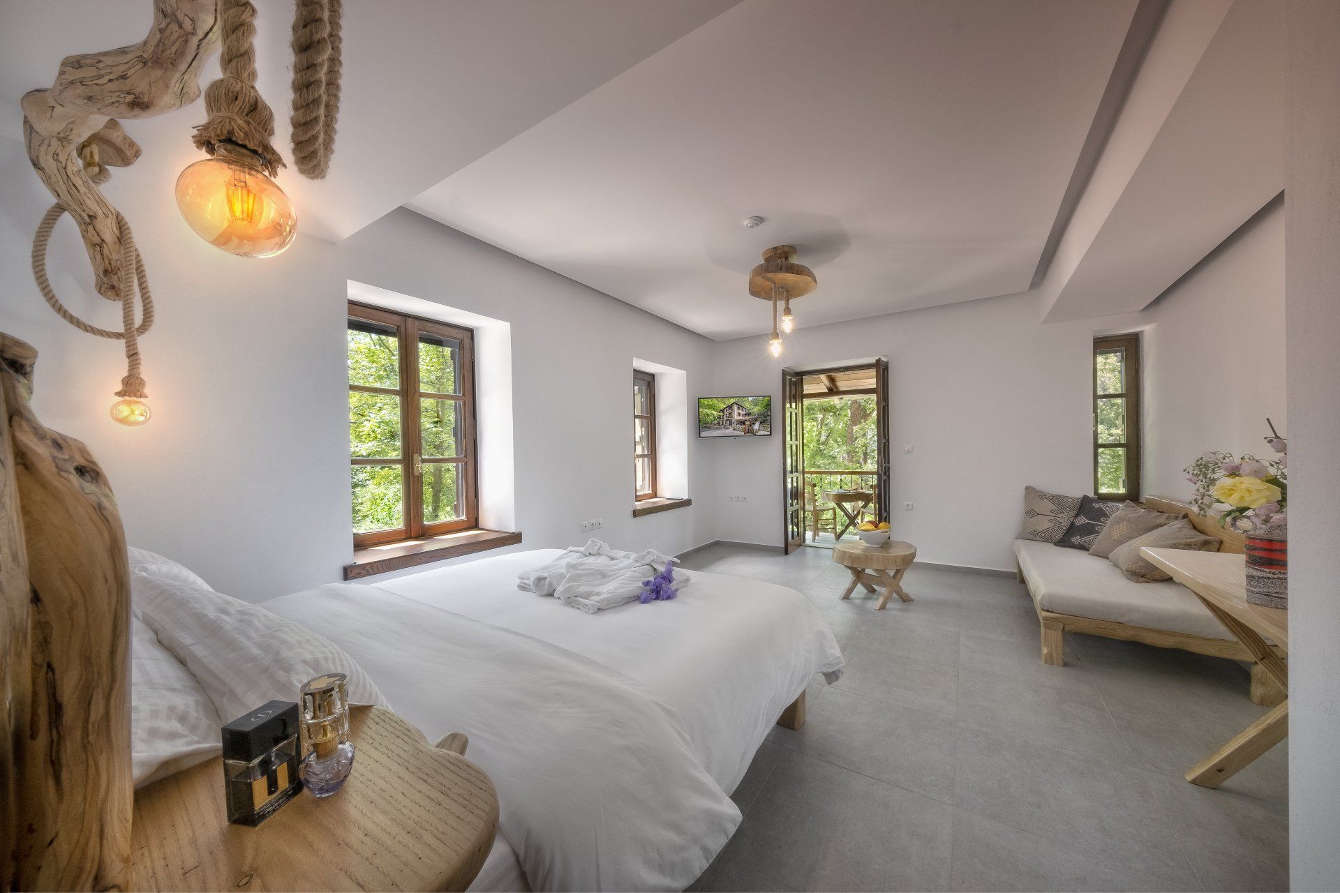 Bedroom with white walls, bed, couch, wooden accents, and a balcony with greenery visible.