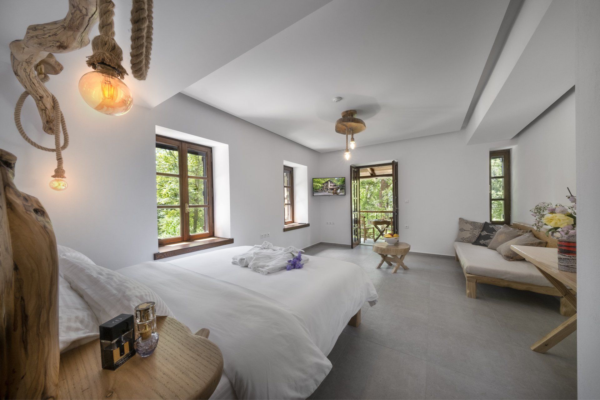 Bedroom with white walls, bed, couch, wooden accents, and a balcony with greenery.