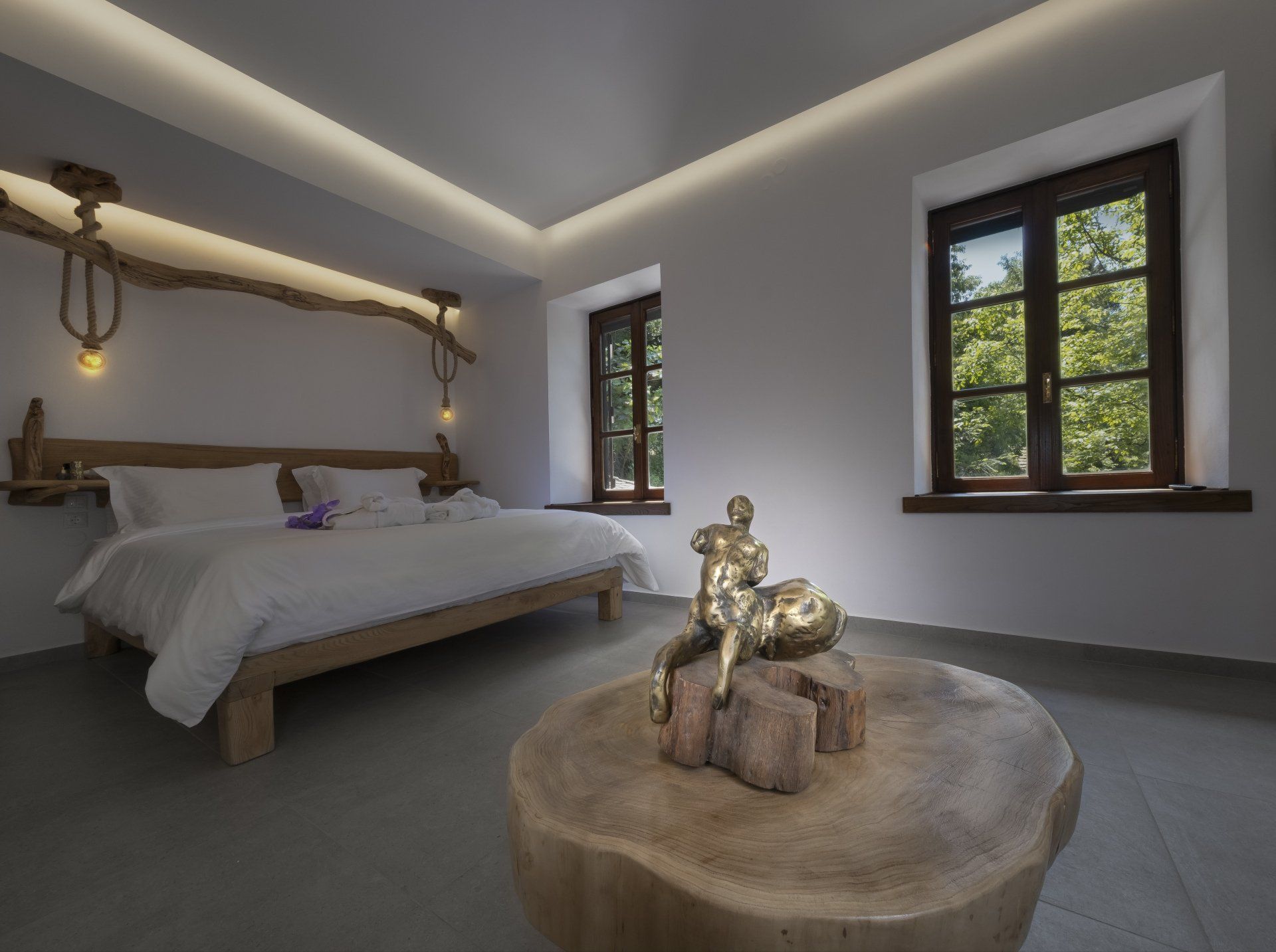 Bedroom with wooden bed, rustic table, golden sculpture, two windows with forest view, and neutral walls.