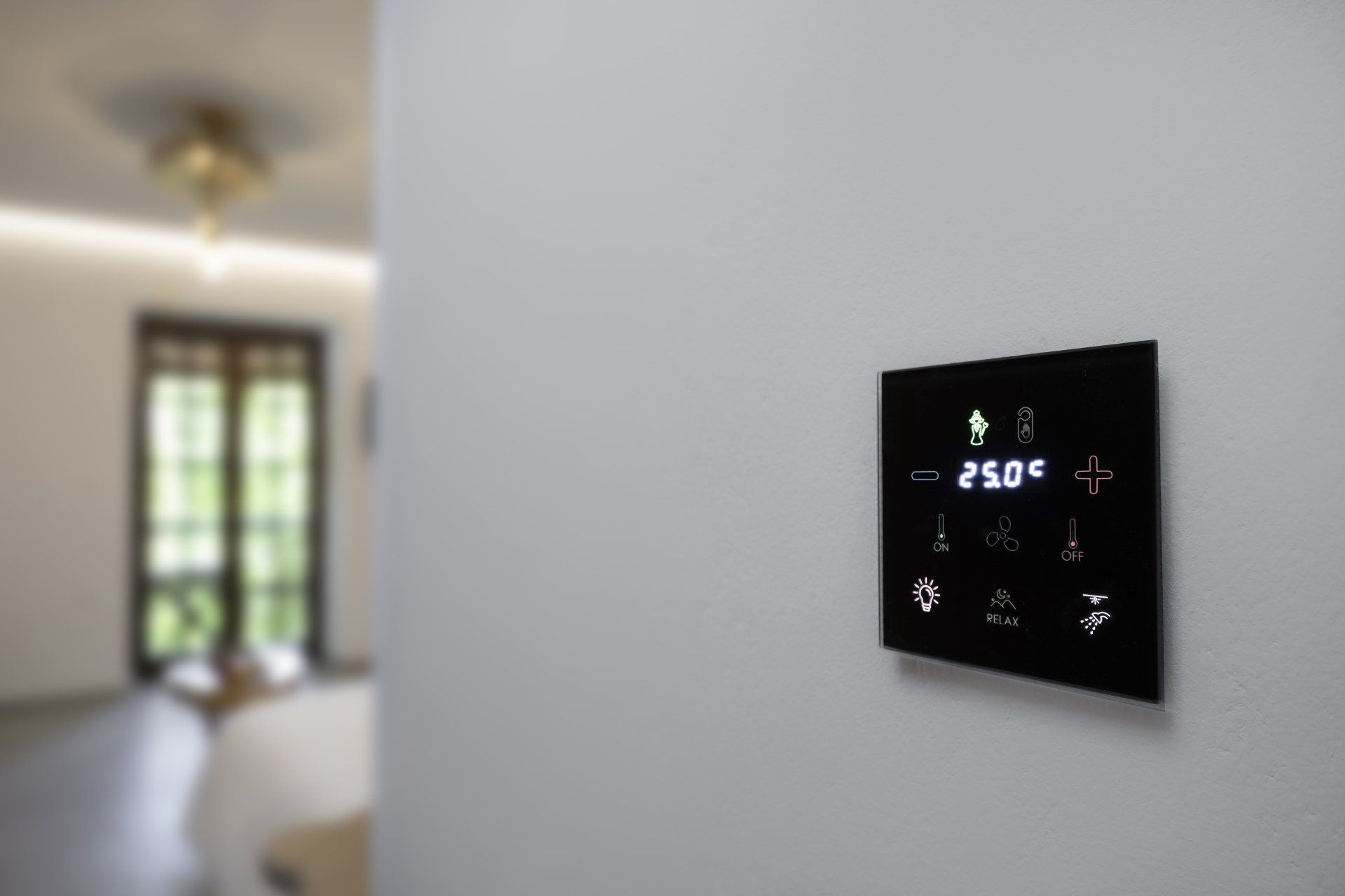 Black touchscreen control panel on a white wall displaying temperature and climate control options. Blurred room background.