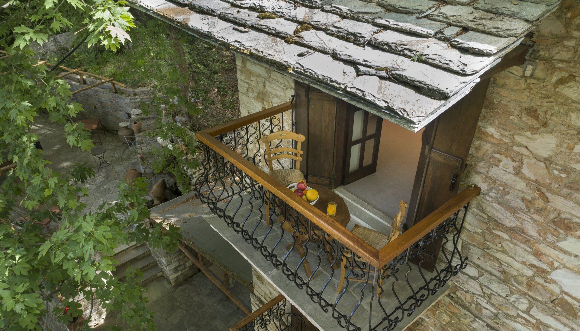 Balcony with a table, chair, and window in a stone building; wooden railing and tile roof.