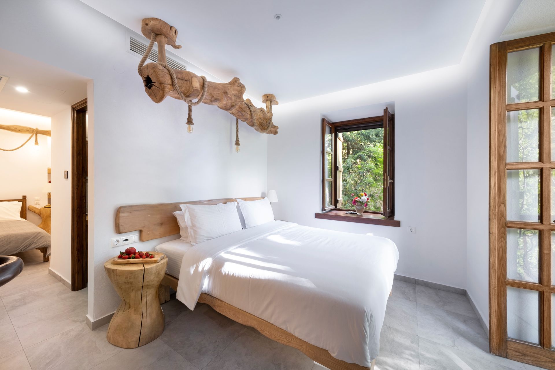 Bedroom with white bedding, wooden accents, and a window overlooking greenery.