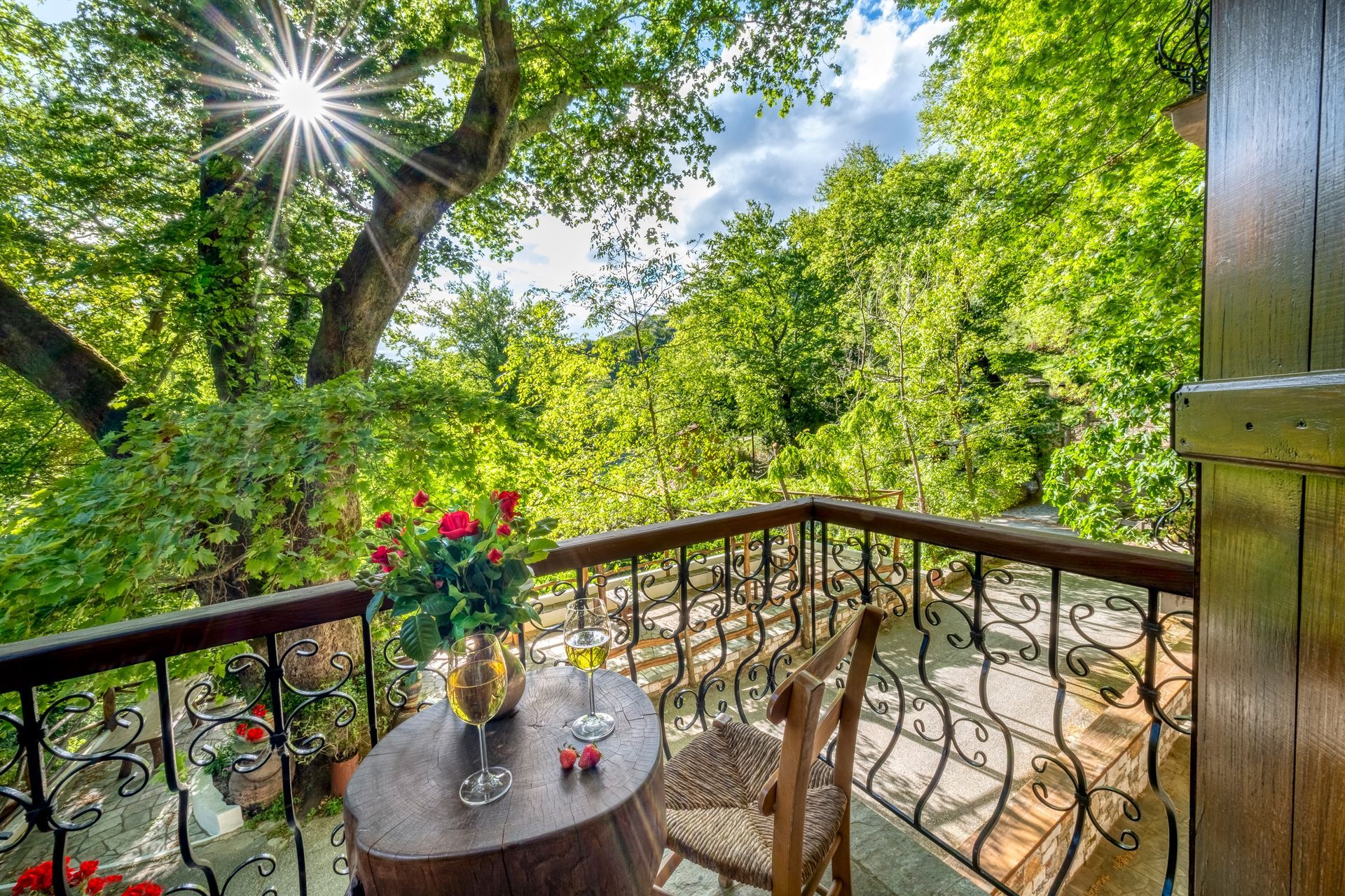 Balcony with table, flowers, and two glasses. Green trees in background, sunny day.