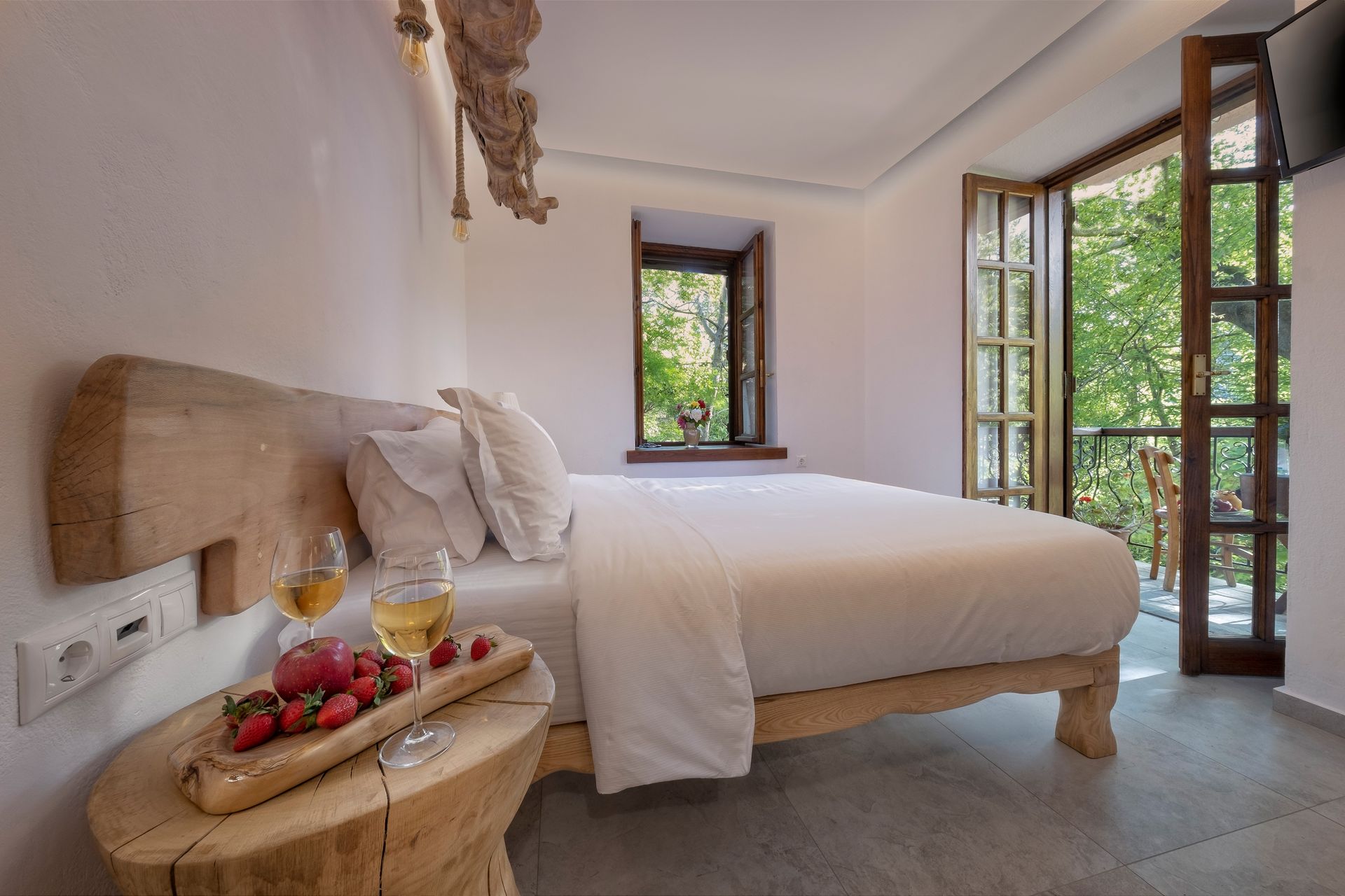 Cozy bedroom with wooden headboard, bed, small table with fruit, open doors with view of trees.