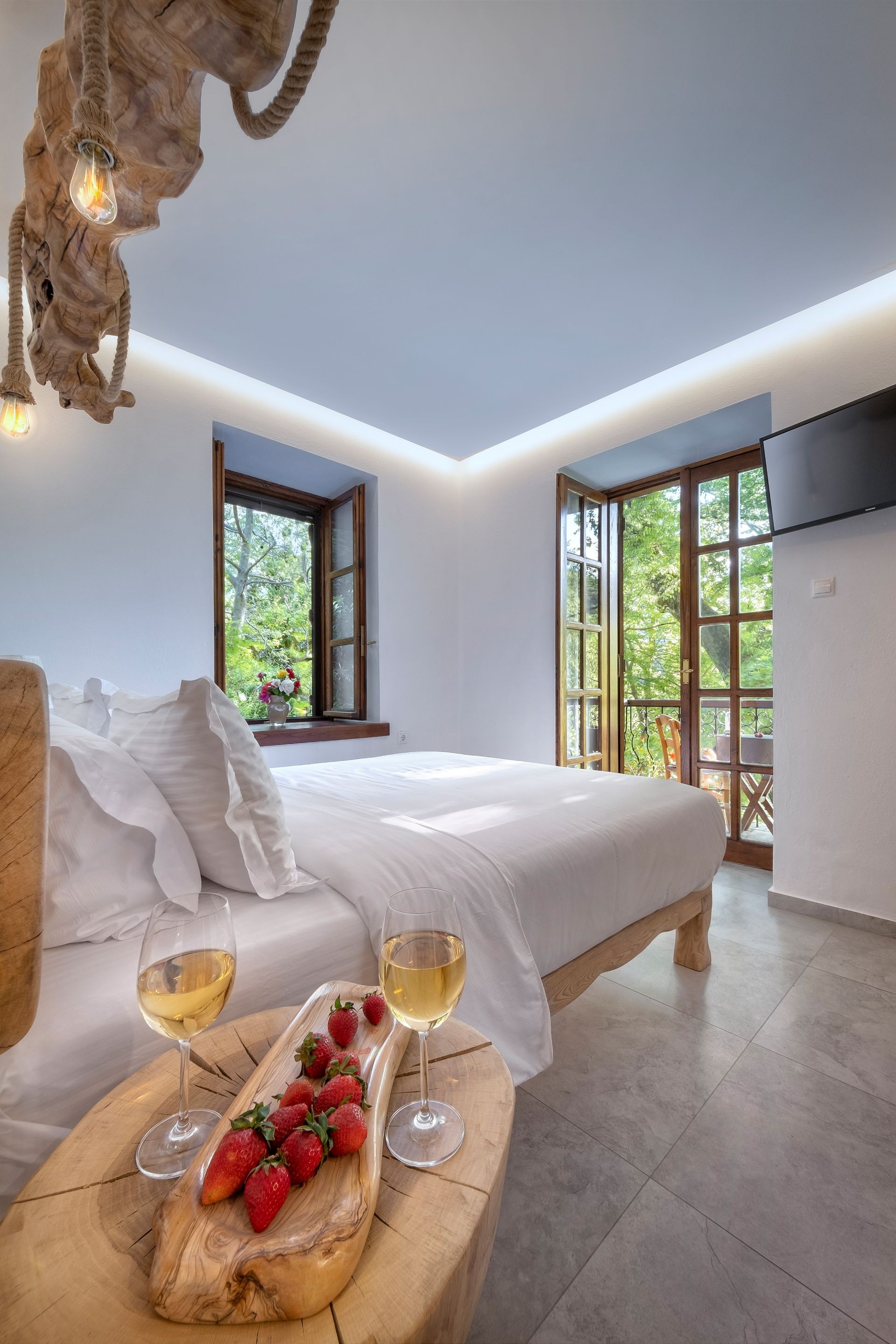 Bedroom with white linens, wooden accent table with wine and strawberries, window, and balcony.