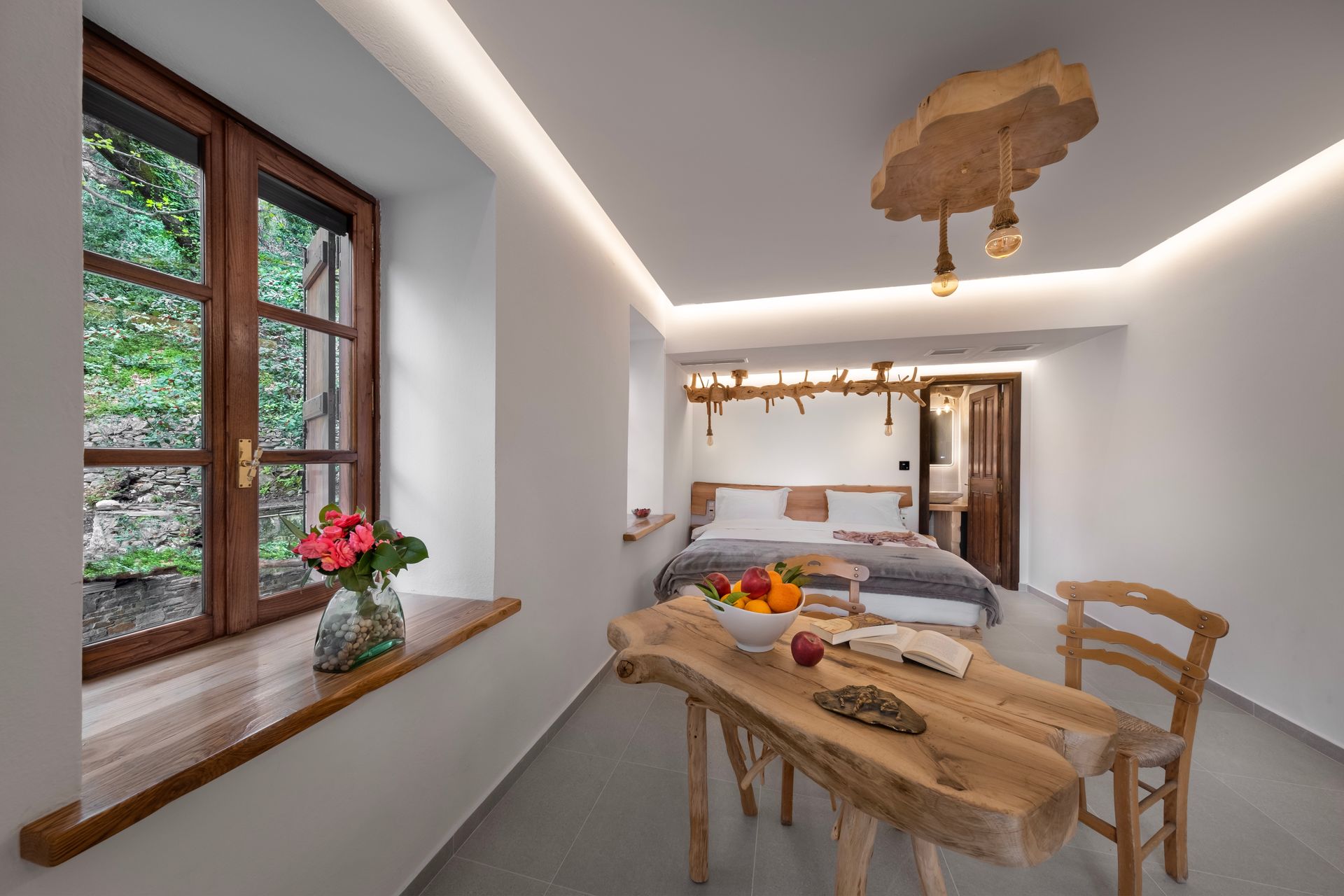 Bedroom with rustic wooden table, window, and bed.  Fruits and books on the table, and hanging lights.