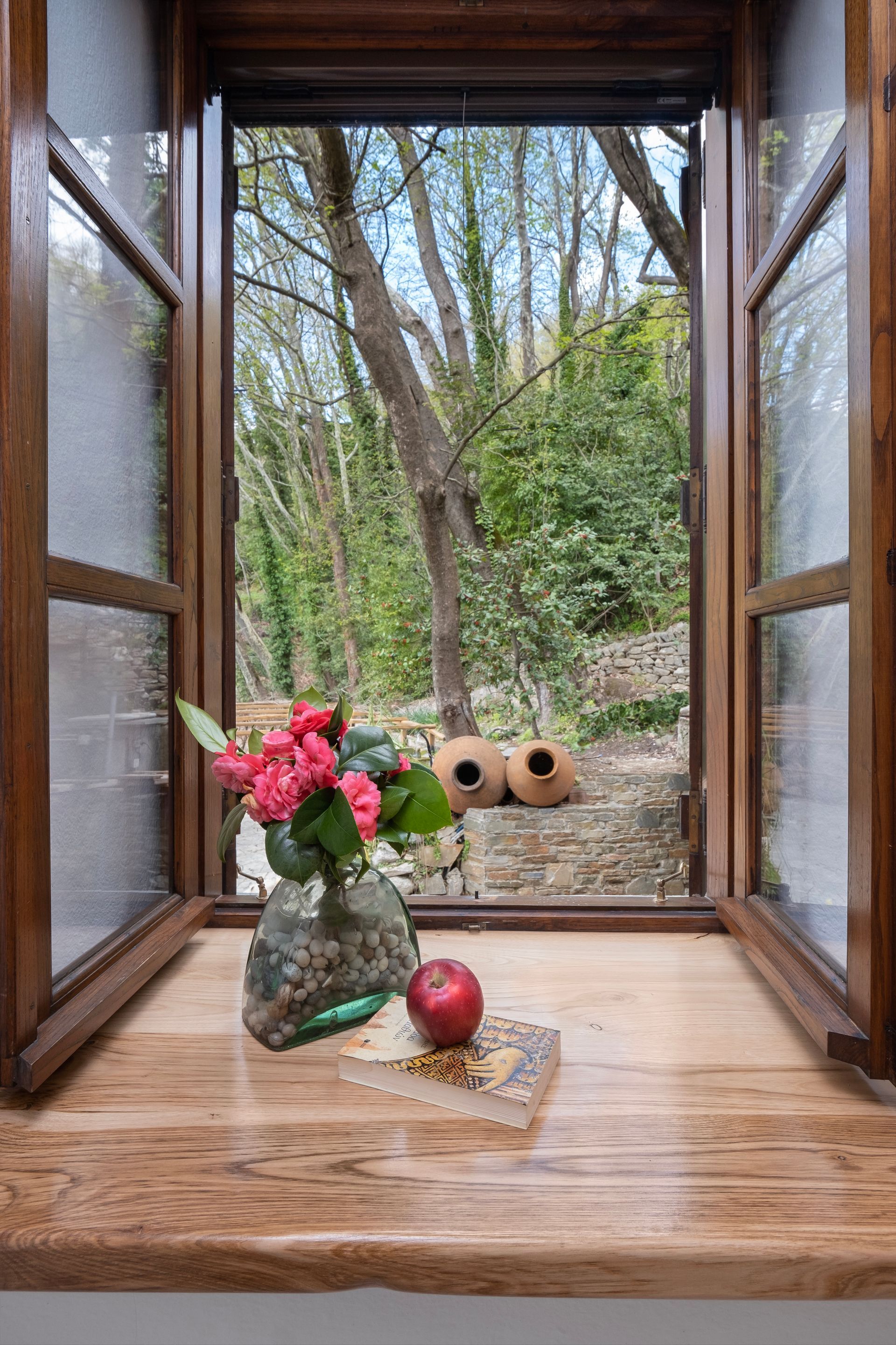 Open wooden window with flowers, apple, and book on the sill, overlooking a garden.