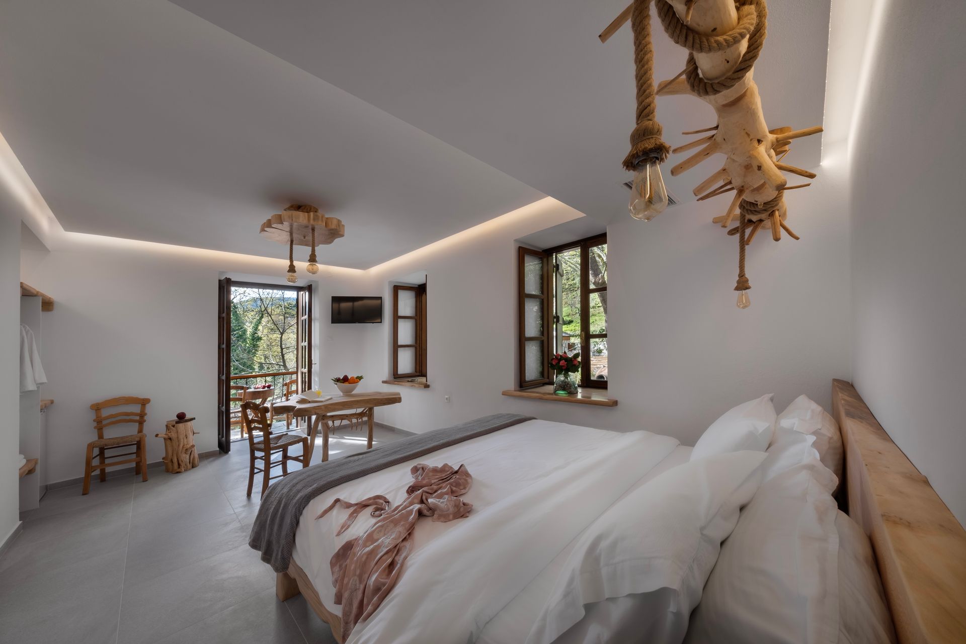 Bedroom with wooden bed, table, balcony, and rustic lighting. White walls, grey floor, natural wood accents.