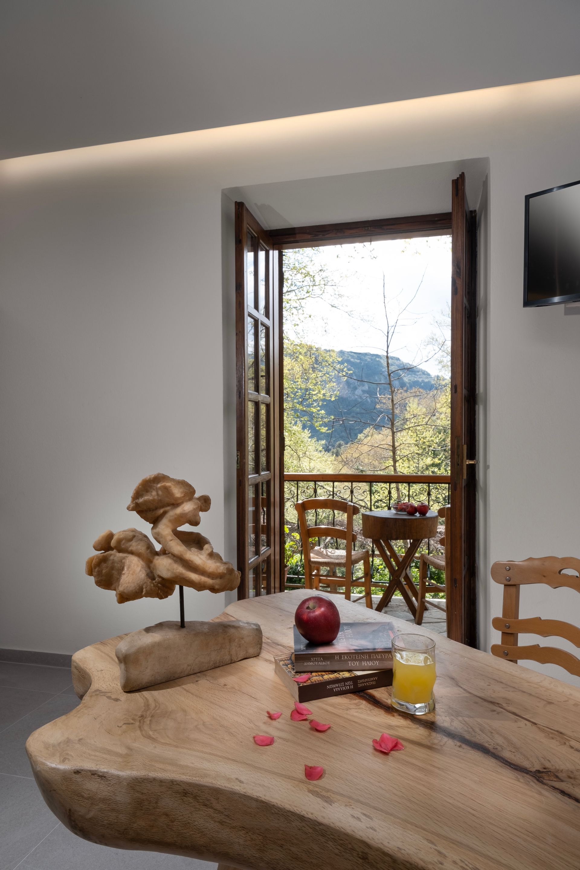 Interior with wooden table, balcony view of trees, fruit, and an artistic sculpture.