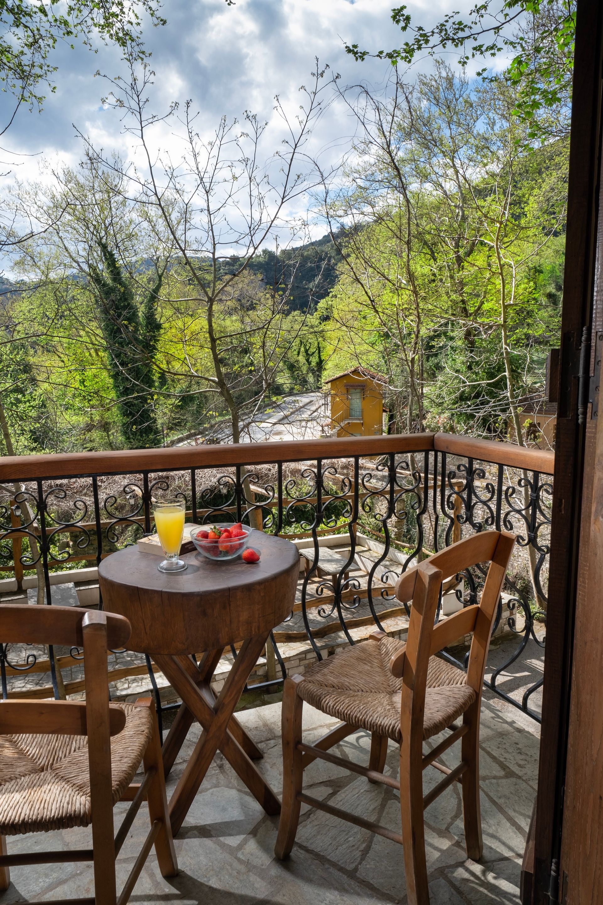 Balcony with table, chairs, juice, and fruit, overlooking a wooded valley and stream.