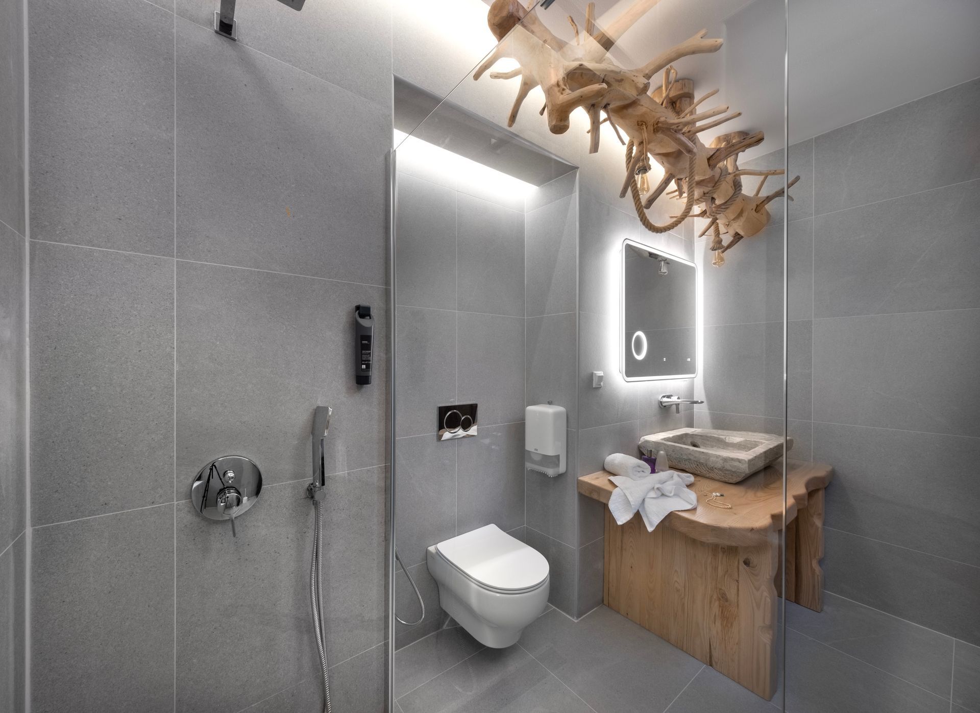 Modern bathroom with gray tile walls, a white toilet, and a wood vanity. A decorative wood sculpture hangs from the ceiling.