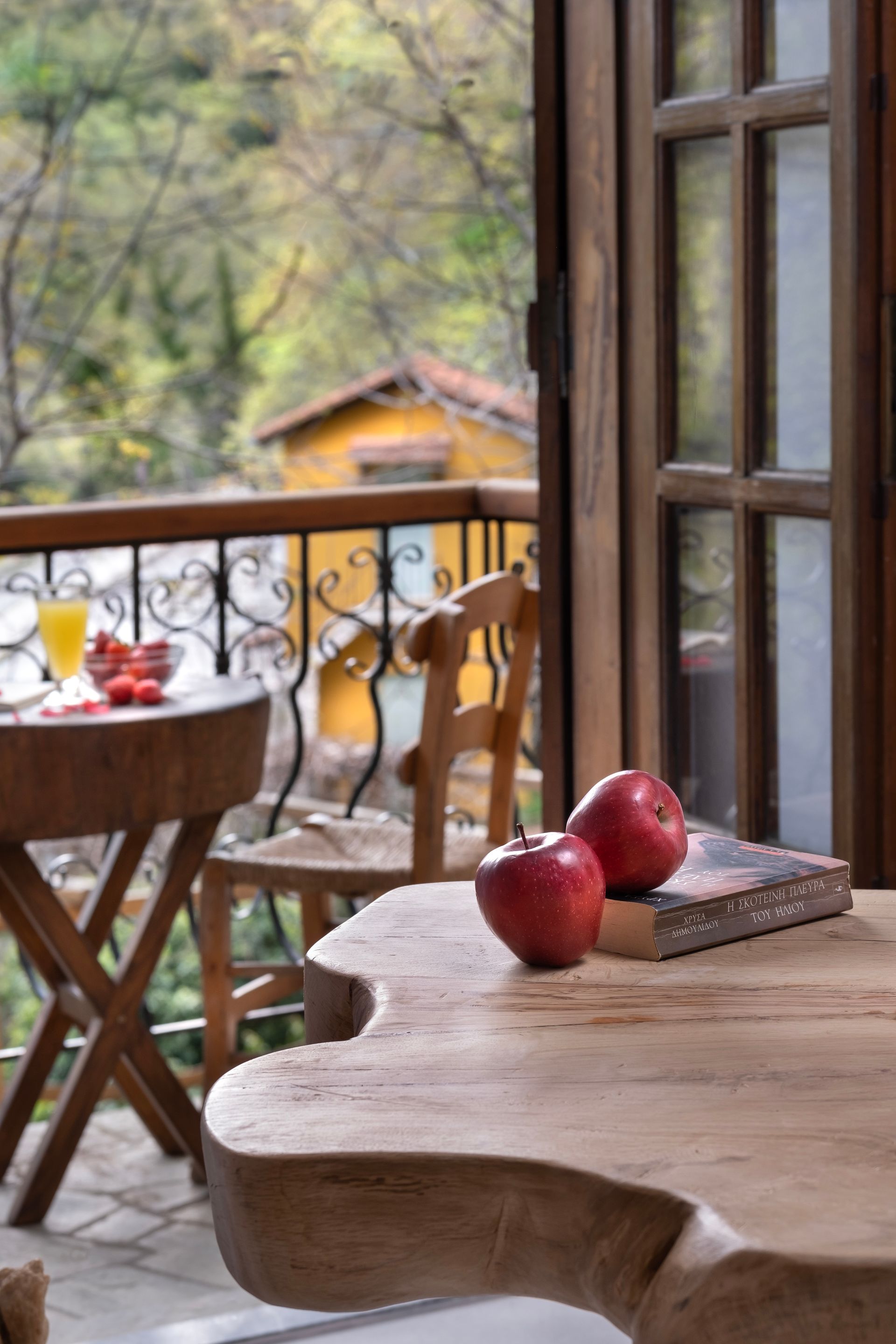 Balcony scene: apples and book on a wooden table. View of forest and building through open doors.