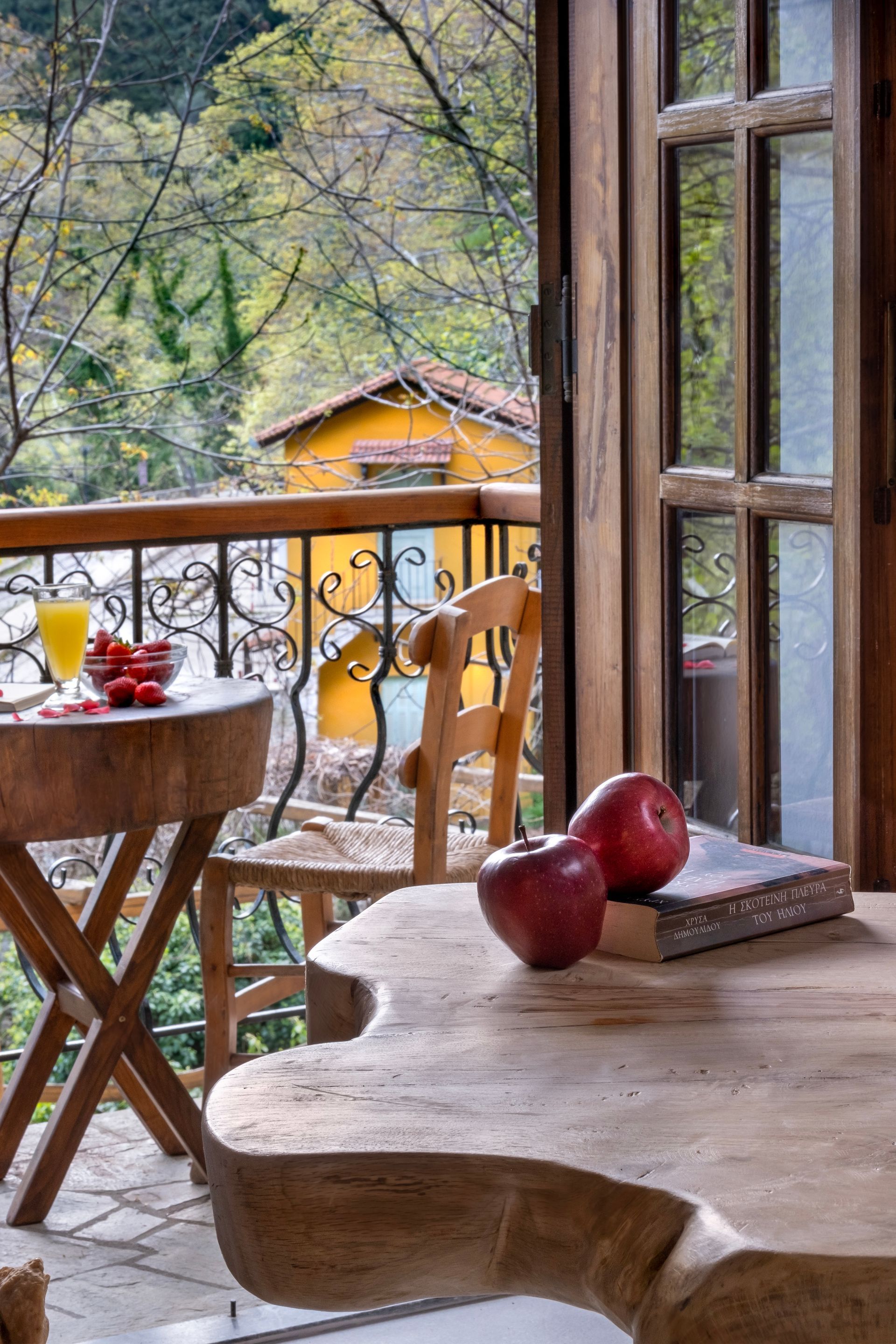 Wooden balcony with table, chair, apples, and open book, overlooking trees and yellow building.