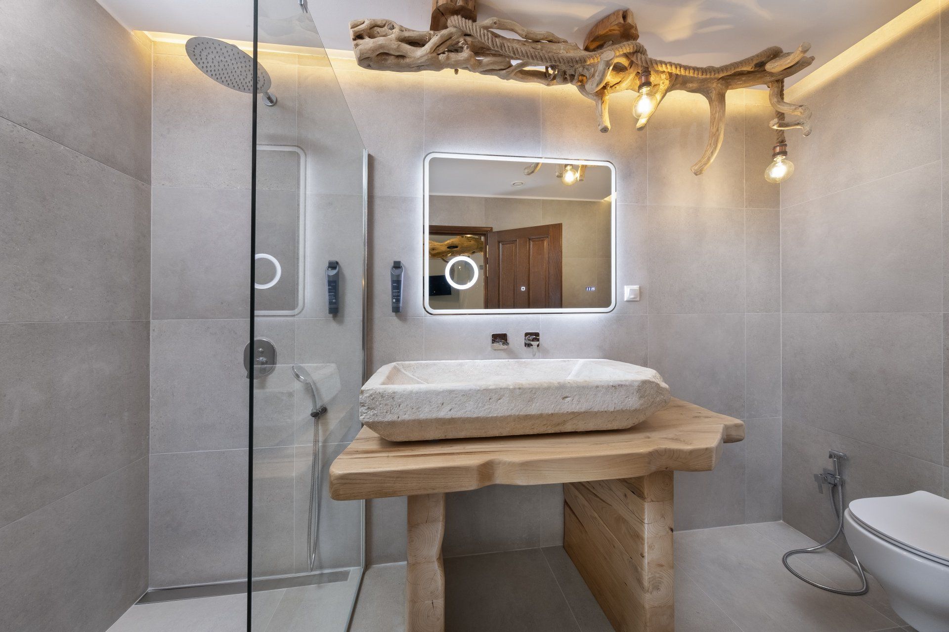 Gray bathroom with a stone sink on a wooden stand, lit mirror, glass shower, and rustic driftwood light fixture.