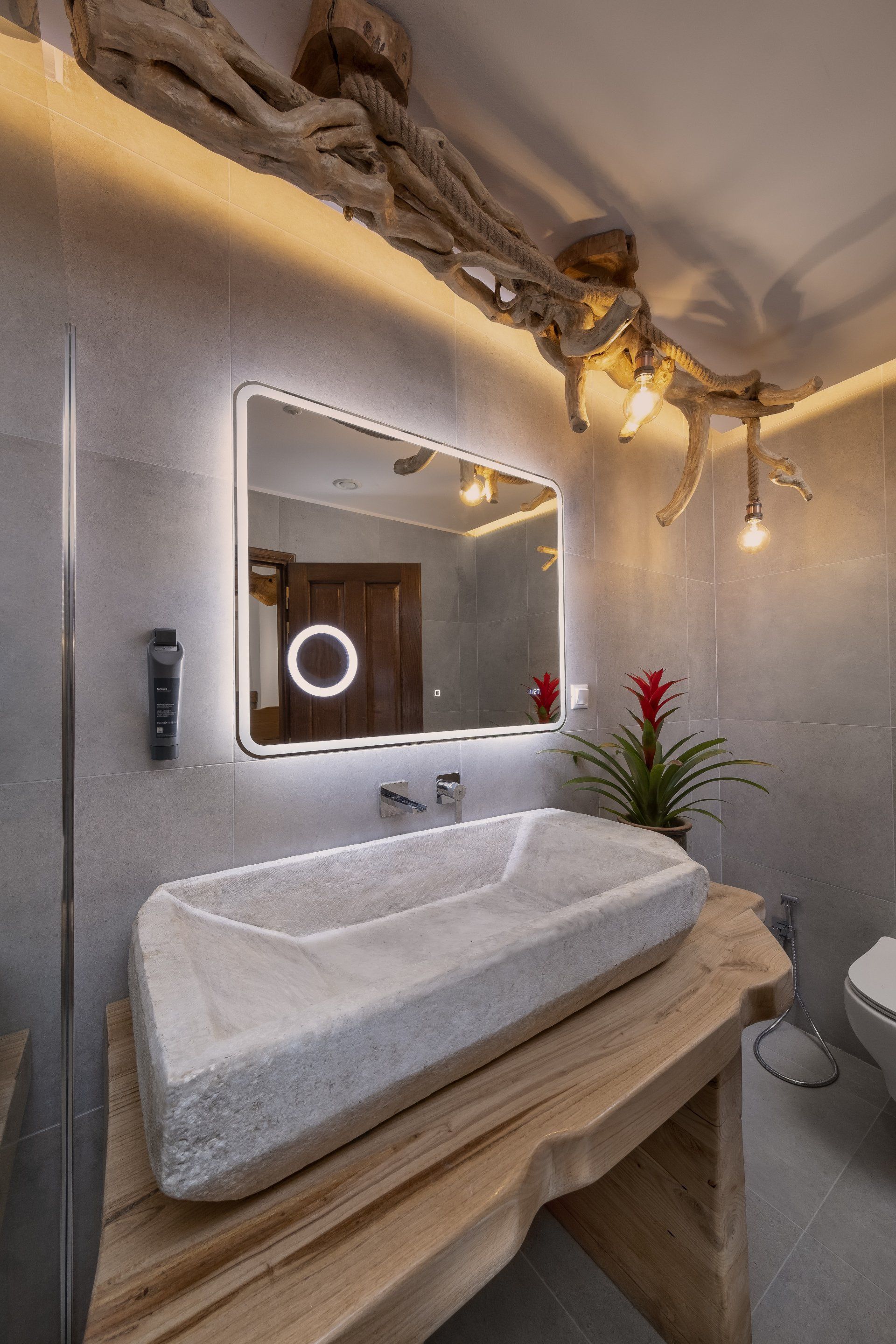 Bathroom with stone sink on a wooden counter, illuminated mirror, and a decorative wooden branch on the ceiling.