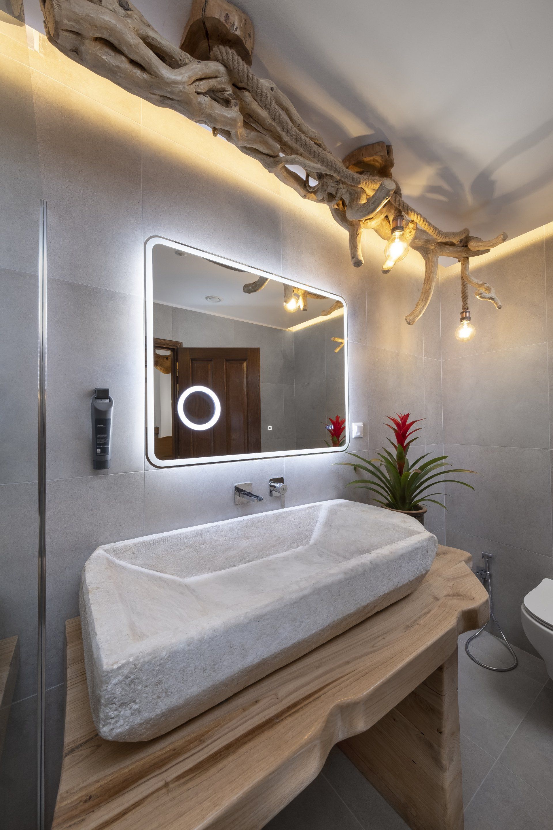 Bathroom with stone sink on wooden counter, lit mirror, and driftwood light fixture.