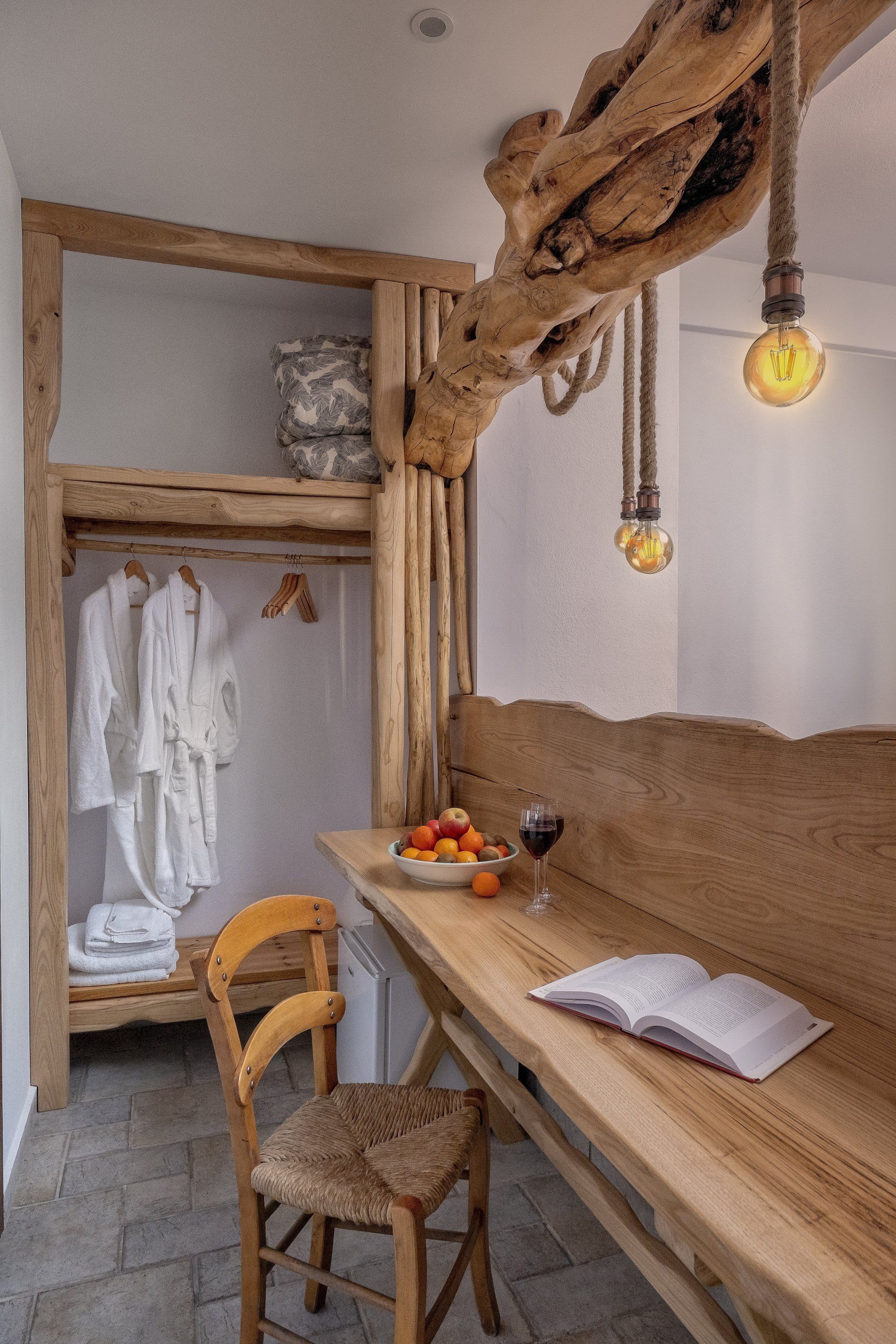 Cozy bedroom corner with wooden beams, closet, desk with fruit and a book, and hanging lights.