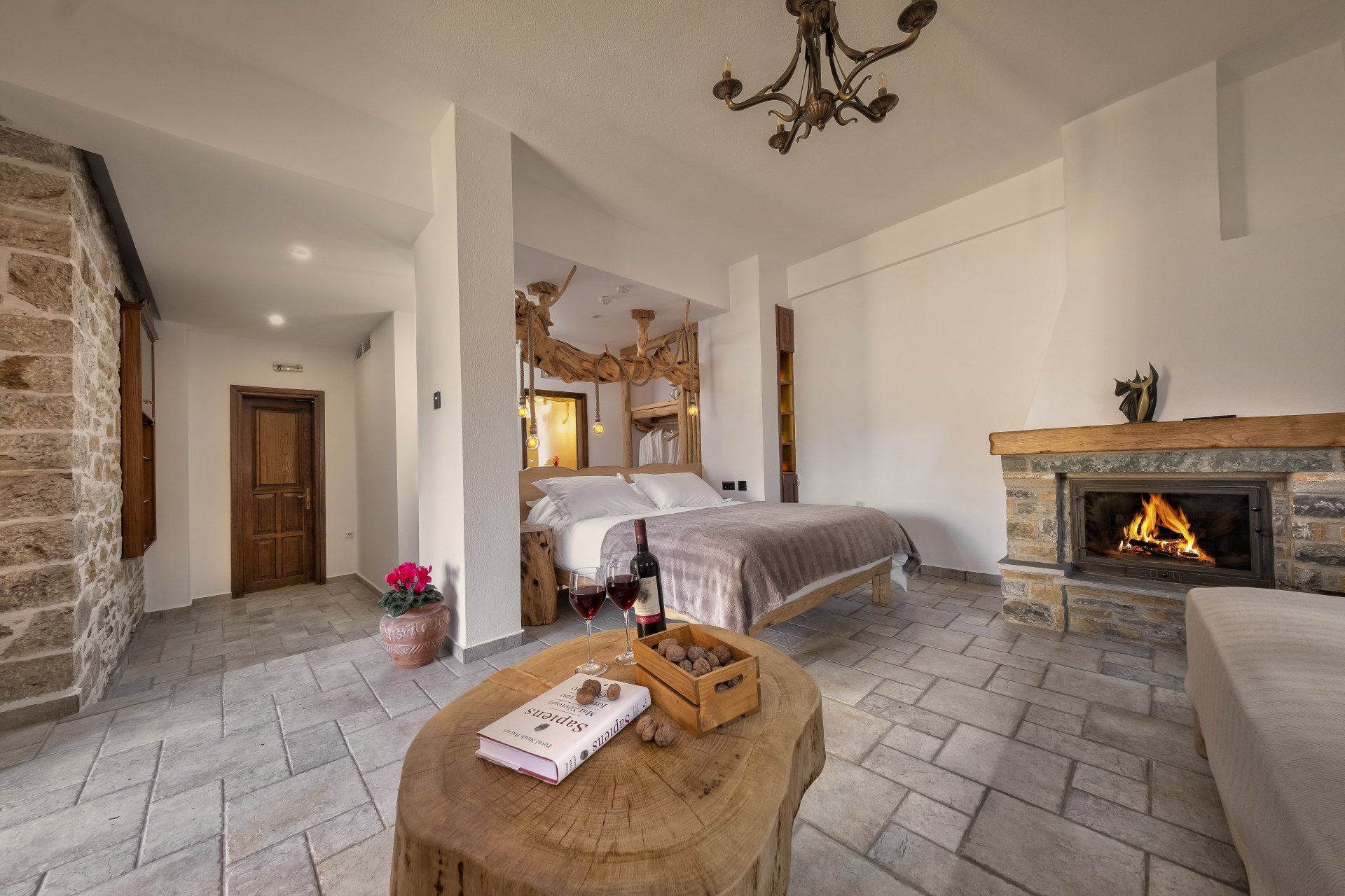Cozy bedroom with fireplace, bed, and rustic wooden table. Warm lighting, stone walls and floor.