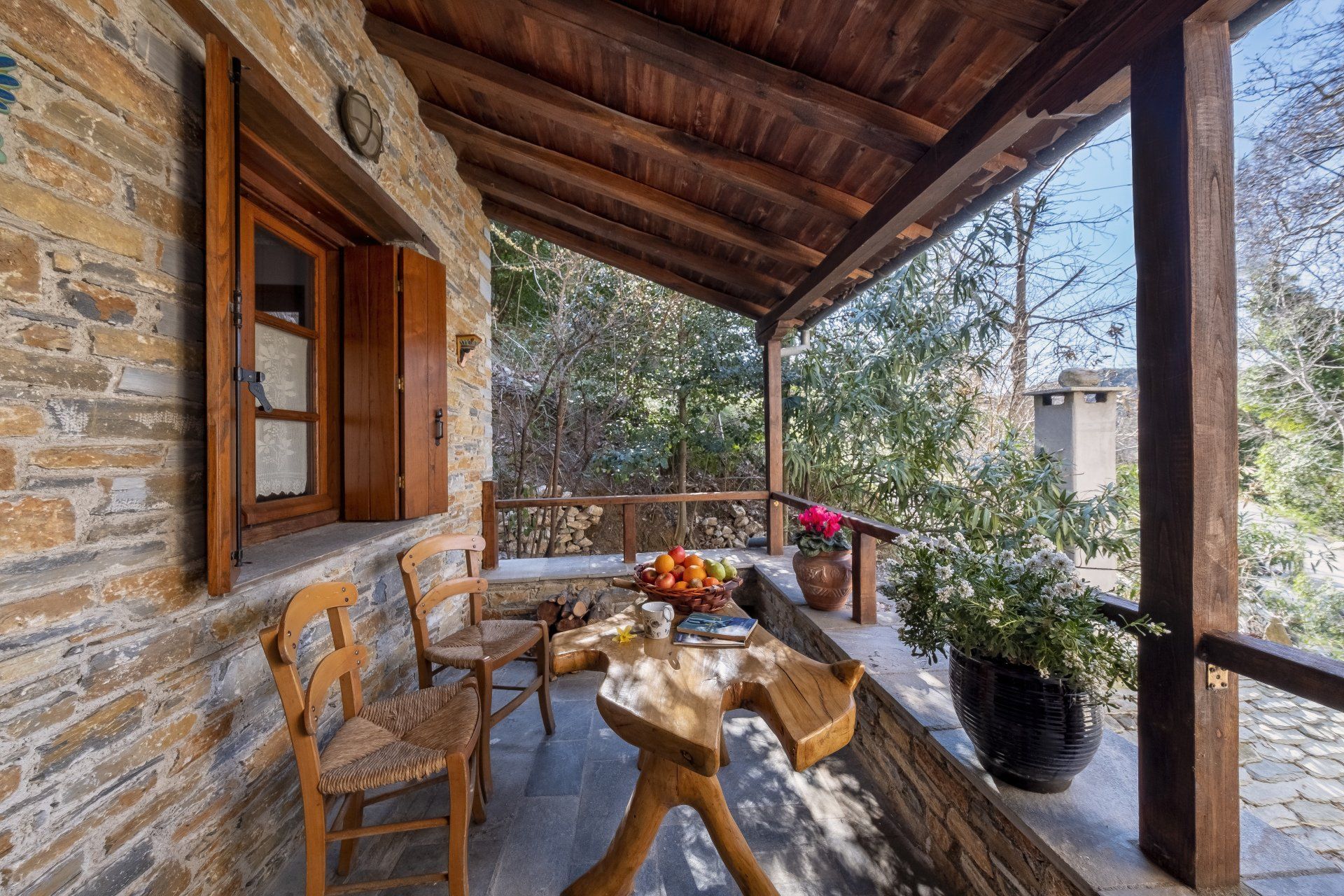 Stone porch with wooden beams, table with fruit, and two chairs. Overlooks trees.