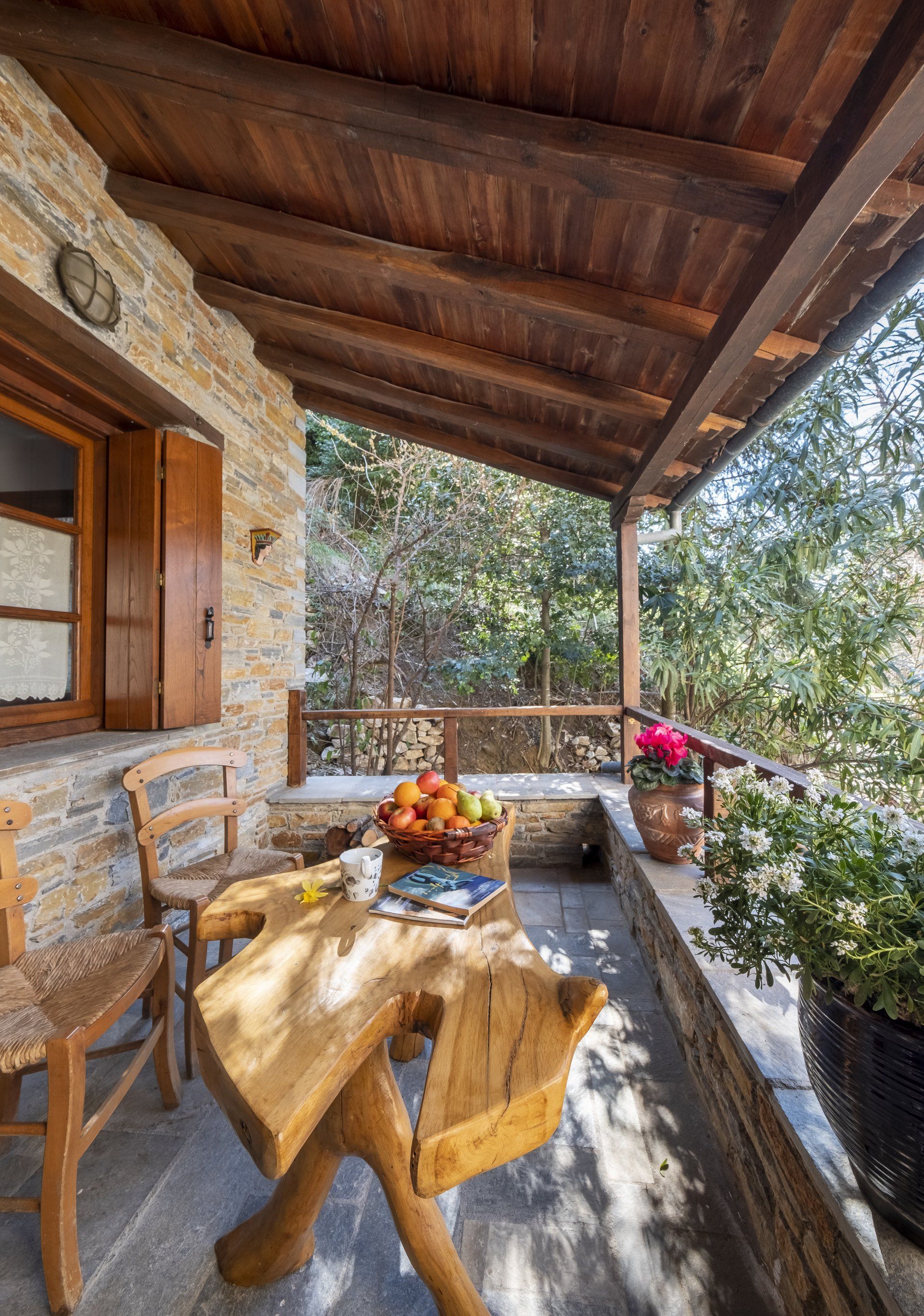 Wooden porch with a rustic table, chairs, and a bowl of fruit overlooking greenery.