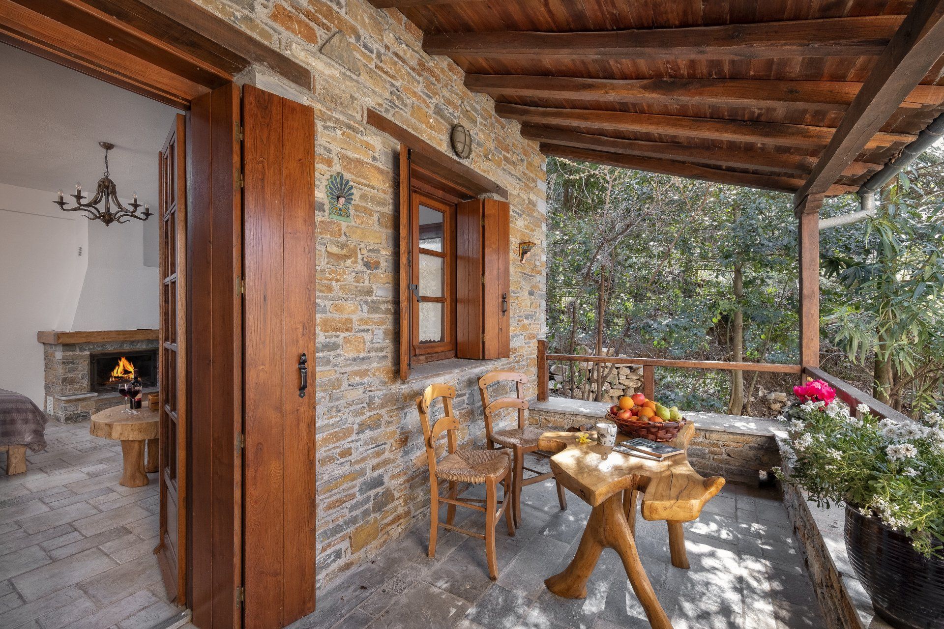 Balcony of a stone cottage with a table, chairs, flowers, and a view of trees.