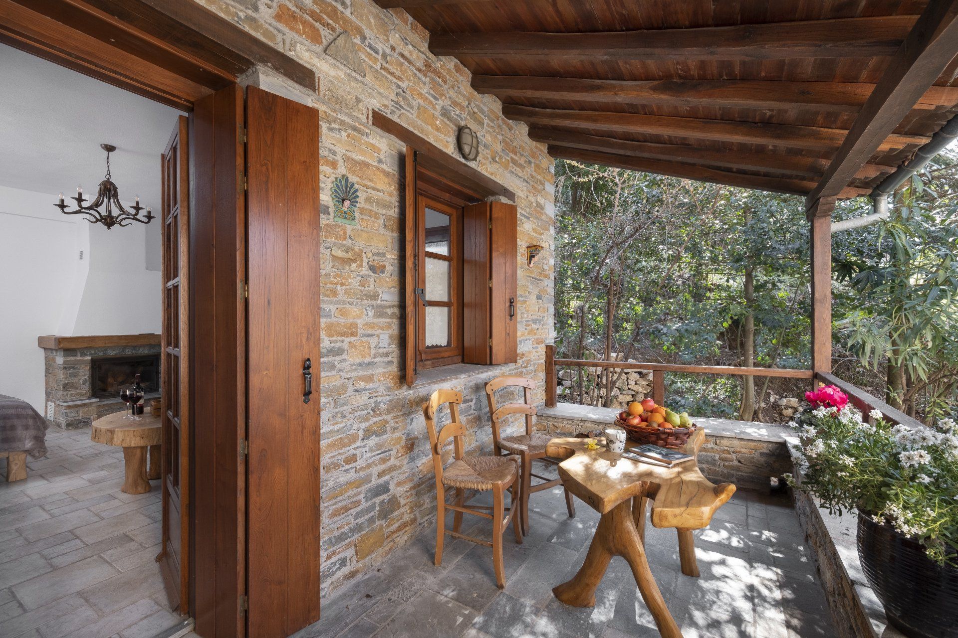 Stone cottage balcony with table, chairs, flowers, and forest view.