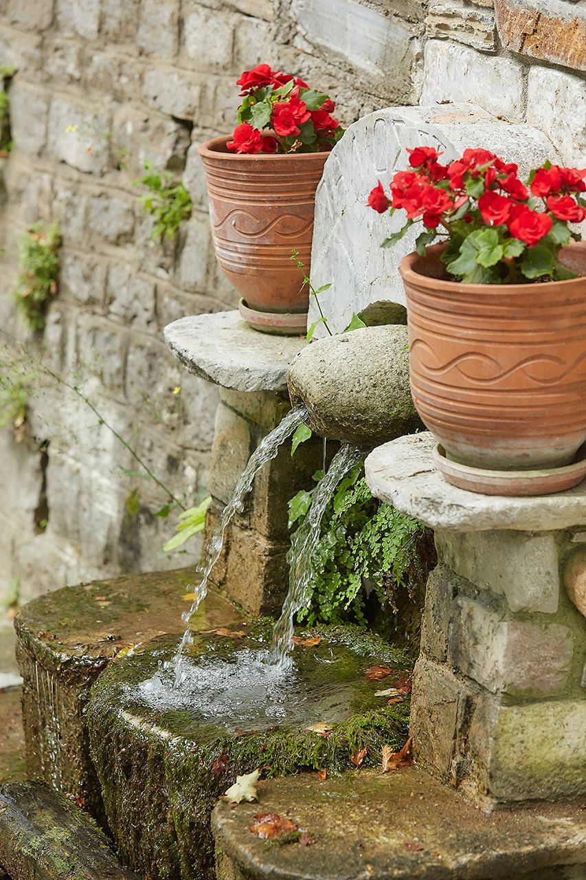 Stone fountain with water spilling into a basin, flanked by terracotta pots of red flowers.
