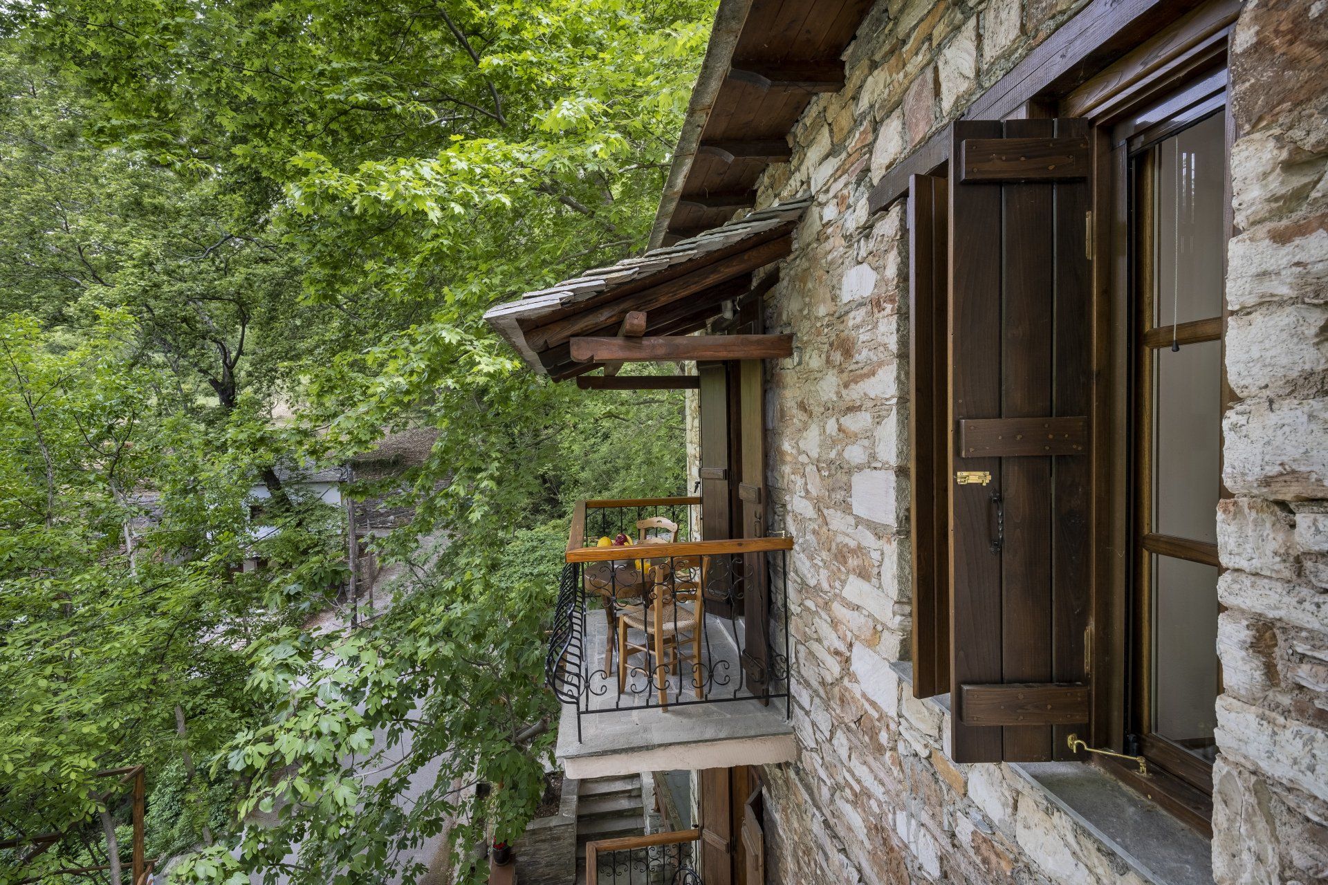 Stone building with a small balcony and open wooden shutters, surrounded by green foliage.
