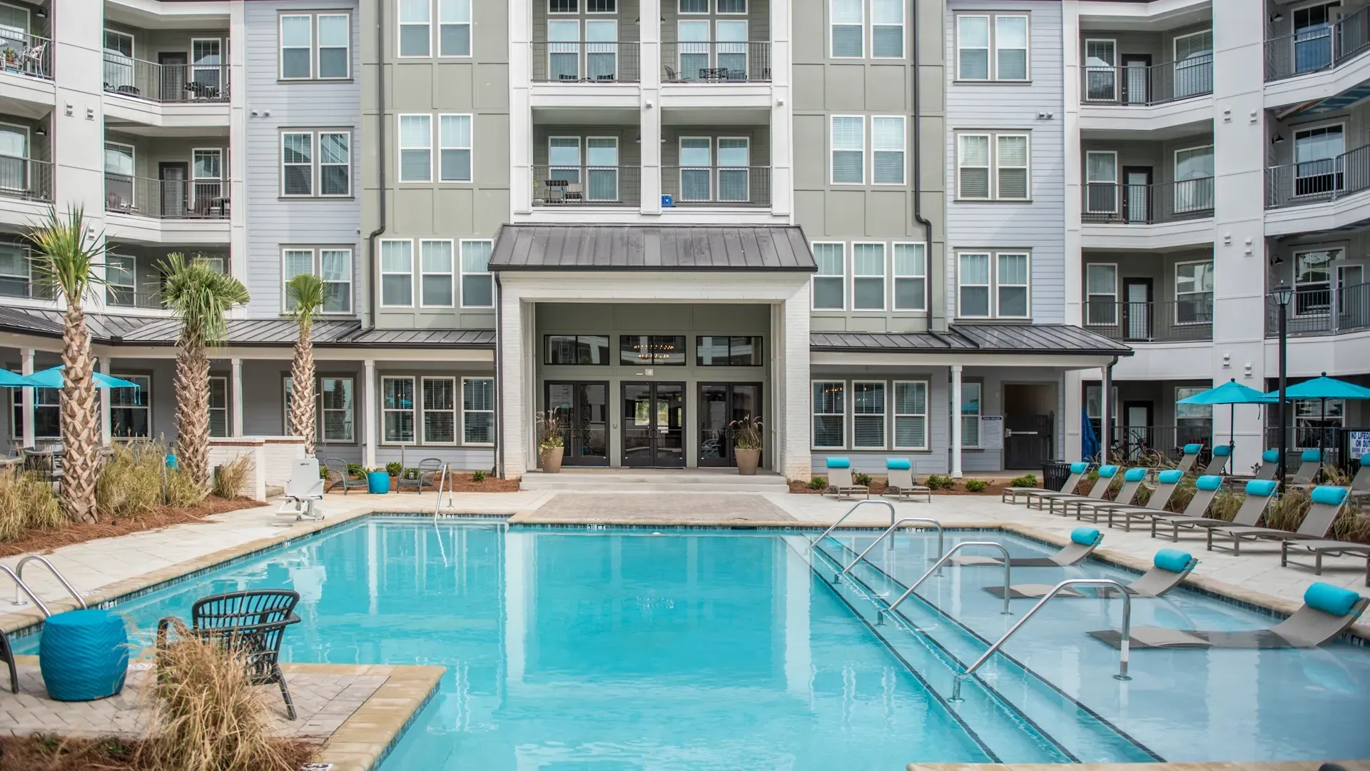 Resort-style swimming pool with lounge chairs and umbrellas in front of apartment buildings.