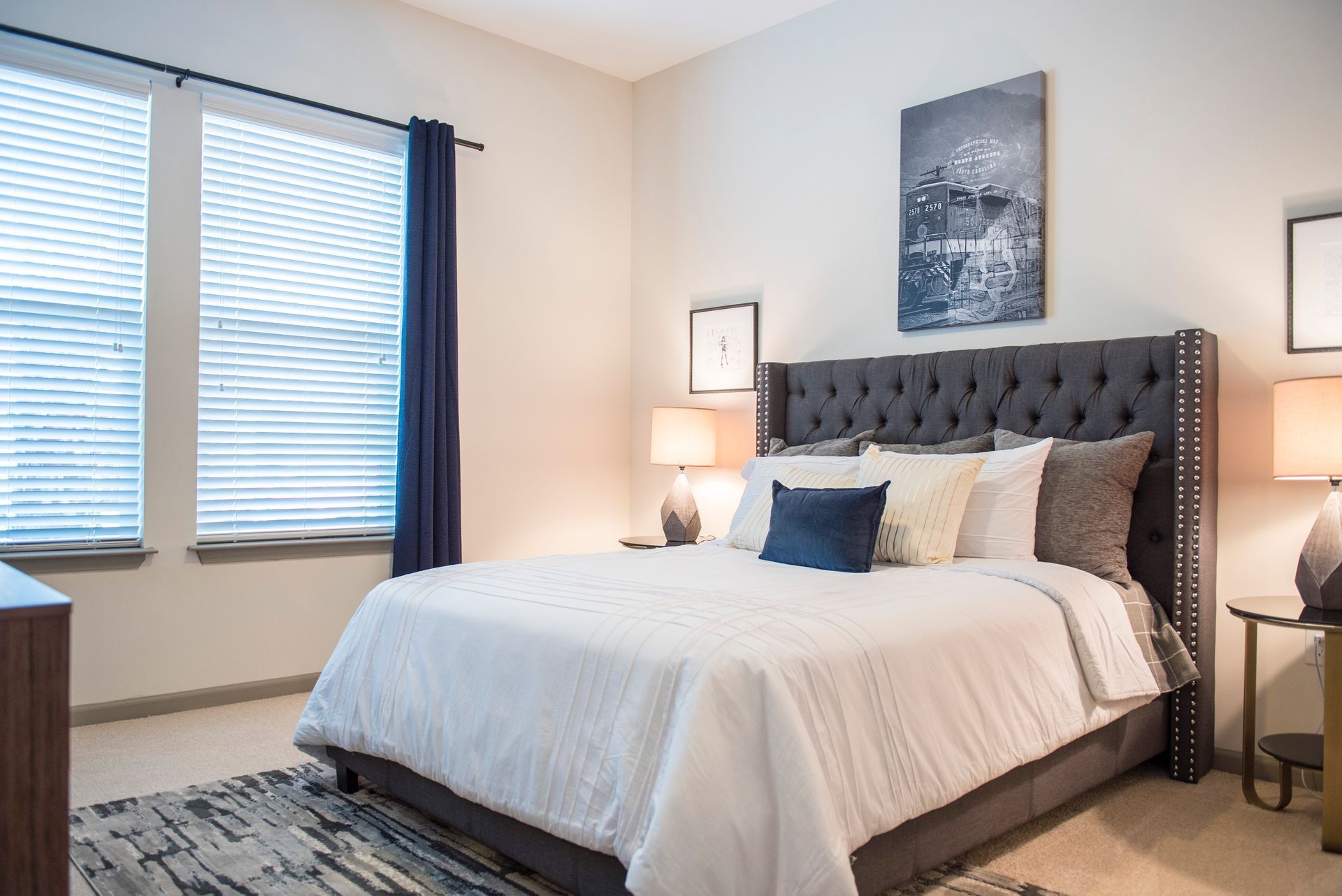 Bright bedroom with a tufted bed, blue accents, and windows with blinds and dark curtains
