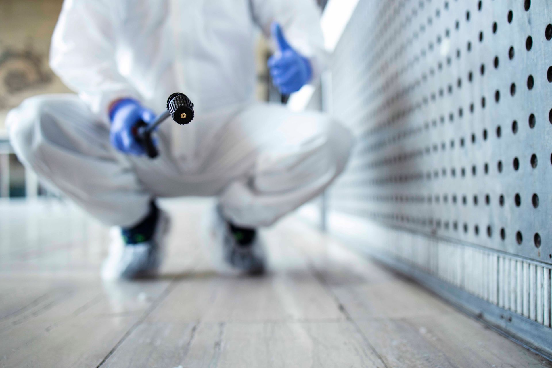 Person in protective suit crouching, holding sprayer, thumb up near a perforated metal wall.