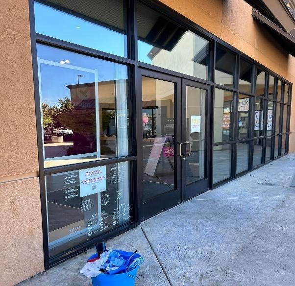 Exterior of a storefront with glass windows and doors; a blue trash can is in front of the window.