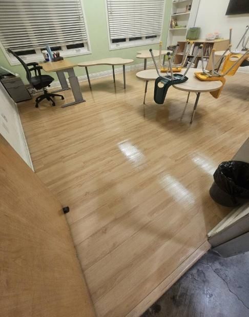 Empty classroom with light wood-look flooring, desks, chairs, and blinds on windows.